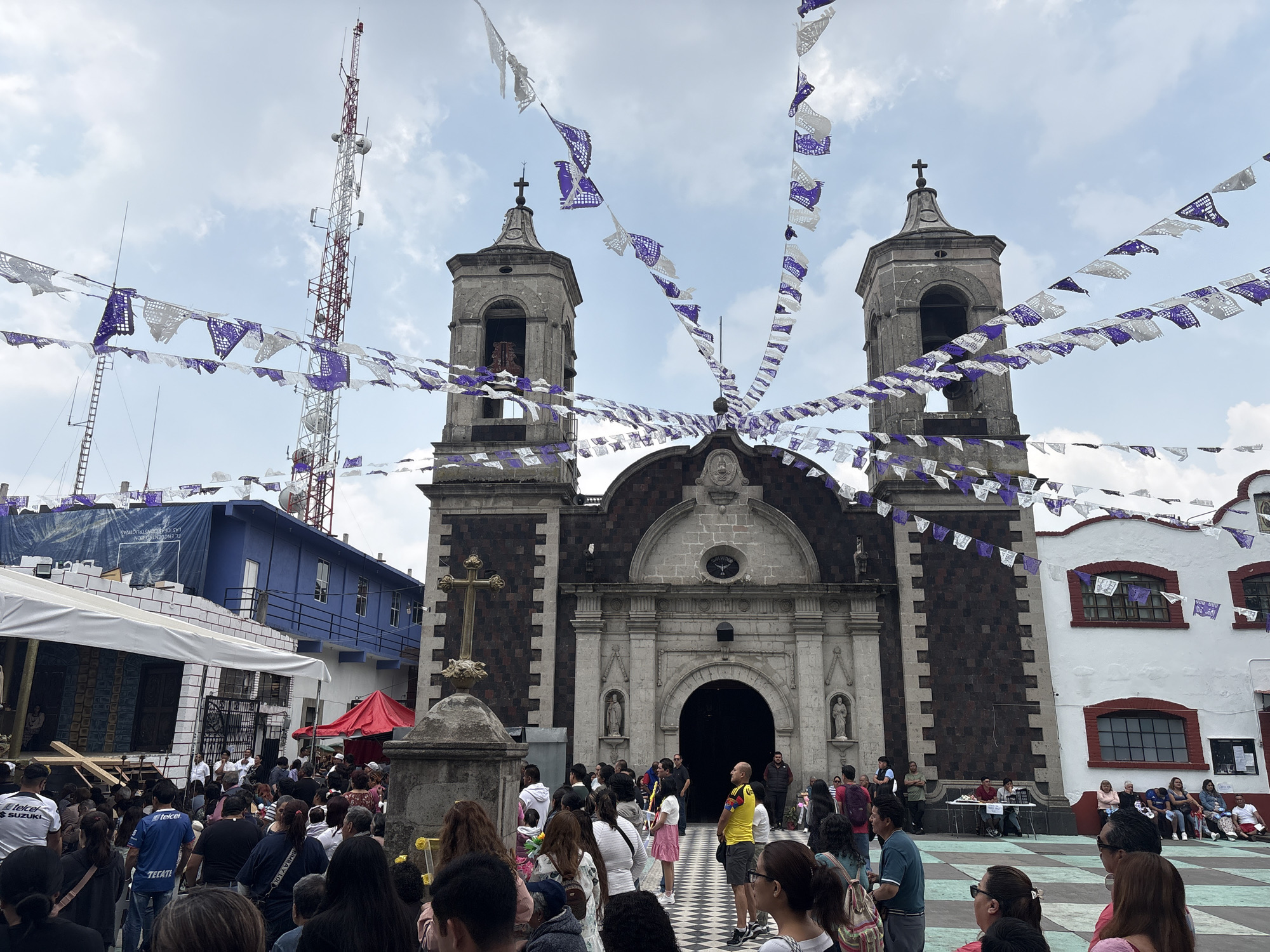 The image depicts a large crowd gathered in front of a church in an outdoor plaza. The church has two towers, each topped with a cross. Papel picado, decorative paper banners, are strung across the plaza. A tall telecommunications tower is visible in the background. People are standing and sitting in groups, some appearing to be in conversation. A cross on a stone pedestal stands to the left of the church entrance. Based on the architectural style of the church and the presence of papel picado, it is likely located in Mexico City, Mexico.