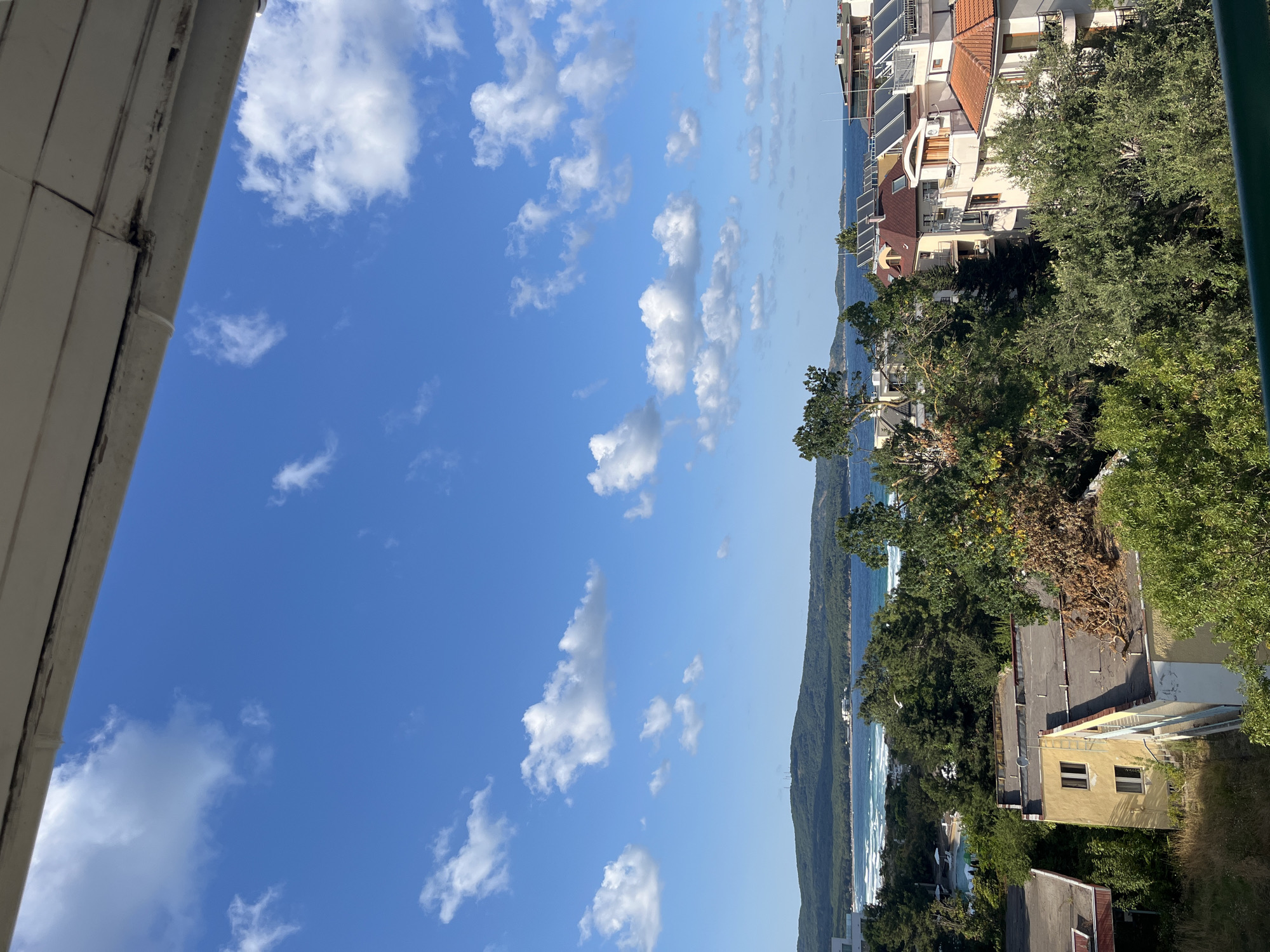 This is a medium-shot, eye-level, outdoor, vertical photograph taken from a balcony or window. The foreground shows the edge of a balcony or overhang with visible signs of wear and tear, including peeling paint and grime. Below the overhang, the scene opens up to a vista of buildings and the sea.

In the mid-ground, a cluster of buildings is visible. Some are residential apartments with tiled roofs and balconies, some of which are equipped with solar panels. One building is painted a pale yellow and appears somewhat neglected, with a ladder leading up to its roof. Dense green trees surround and partially obscure the buildings, with some dry, brown foliage visible on a few branches.

Beyond the buildings and trees, a wide expanse of the sea stretches across the horizon. The water is a deep blue, with white waves breaking near the shore, suggesting a beach or coastline. In the distance, a green, hilly landscape forms the backdrop to the sea, with a few structures and radio towers visible on the highest point. The sky is a vibrant blue, dotted with fluffy white clouds. The overall impression is of a coastal town with a mix of residential development and natural scenery. The location appears to be Primorsko, Bulgaria, based on the landscape and architecture. There are no people or animals visible in the photograph.