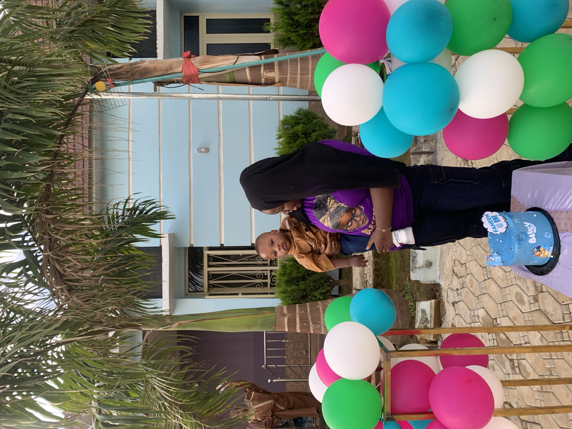 This detailed image captures an outdoor birthday celebration in a residential area of Jos, Nigeria. In the immediate foreground, a woman with dark skin and a black head covering (hijab) is visible, wearing a purple short-sleeved t-shirt with a graphic print featuring a woman's face and text that appears to read "BLESSED." She wears dark blue trousers and has a henna-like design on her right wrist. The woman cradles an infant in her left arm, supporting its back with her right hand while holding a white baby bottle. The baby, also with dark skin, wears a light brown collared long-sleeved shirt and blue trousers, looking directly at the camera with a neutral expression.
To their right, a small table covered with a light purple tablecloth holds a round, two-tier blue birthday cake on a black stand. The cake is decorated with "Bluey" characters around its base and features a white cloud-shaped topper with blue text that reads "FAHEEM @ ONE." Surrounding the table and extending across the foreground are numerous inflated balloons in hot pink, bright green, white, and light blue (teal), some arranged in clusters and supported by gold-colored metal frames.
The background features two houses. On the right, a light blue house with horizontal white trim lines is visible, topped with brown roof tiles and featuring windows with white frames and security bars. A wall-mounted light fixture is present on its facade. On the left, a partially visible purple house with a brown roof and a metal railing on a porch or balcony can be seen. Standing between these structures is a prominent palm tree with green fronds, its trunk wrapped with red fabric ties and a green pipe, possibly supporting an antenna-like object (a yellow disk and a metal grate) higher up. Green shrubbery and low bushes are visible at the base of the houses. On the far left, a person in a long brown garment and head covering is partially visible near the purple house's railing, with their arms slightly raised. The ground is a paved or tiled outdoor area with some scattered dirt and concrete elements.
