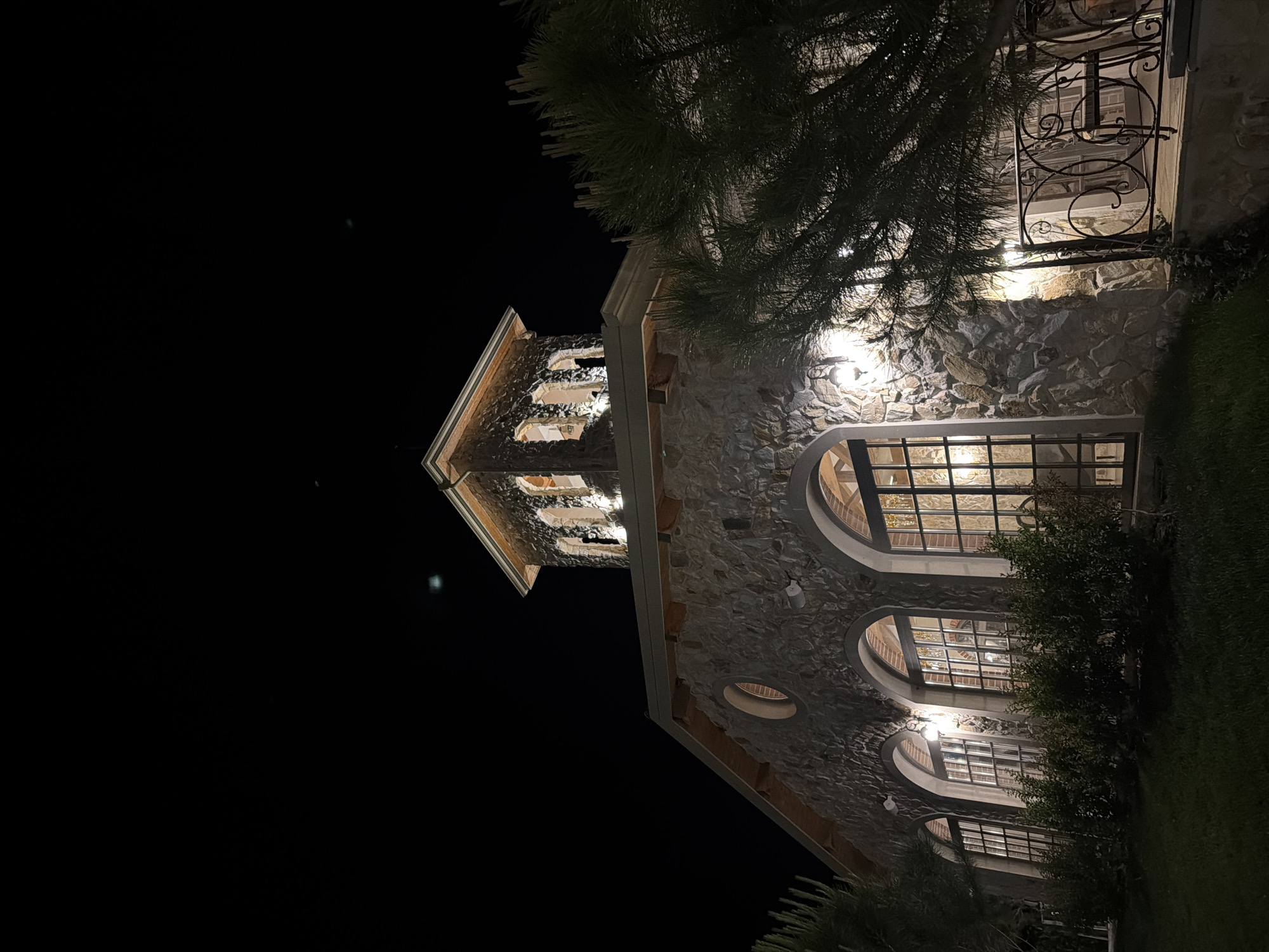 The image shows a stone building at night, illuminated by external lights. The building has a rustic appearance, with a tower topped by a pitched roof. Large arched windows with multiple panes are visible on the facade, and a circular window is present on the upper level. A wrought-iron balcony with a chair is visible on the right side of the building, partially obscured by a pine tree. The ground in front of the building is covered with grass. The location is identified as the town of Kocherinovo, Bulgaria, specifically the St. George the Conqueror winery.