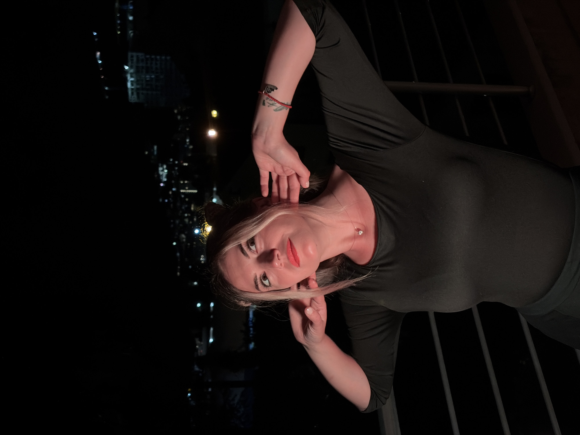 A woman with blonde hair styled in two buns stands on a balcony overlooking a city at night. She is wearing a dark, long-sleeved shirt and has her hands near her ears. A small necklace with a pendant is visible around her neck. On her left forearm, a tattoo of a winged creature is visible, along with a red string bracelet. In the background, the lights of Budva, Montenegro, are visible, with some buildings and cranes indicating construction. The overall lighting suggests it is nighttime.