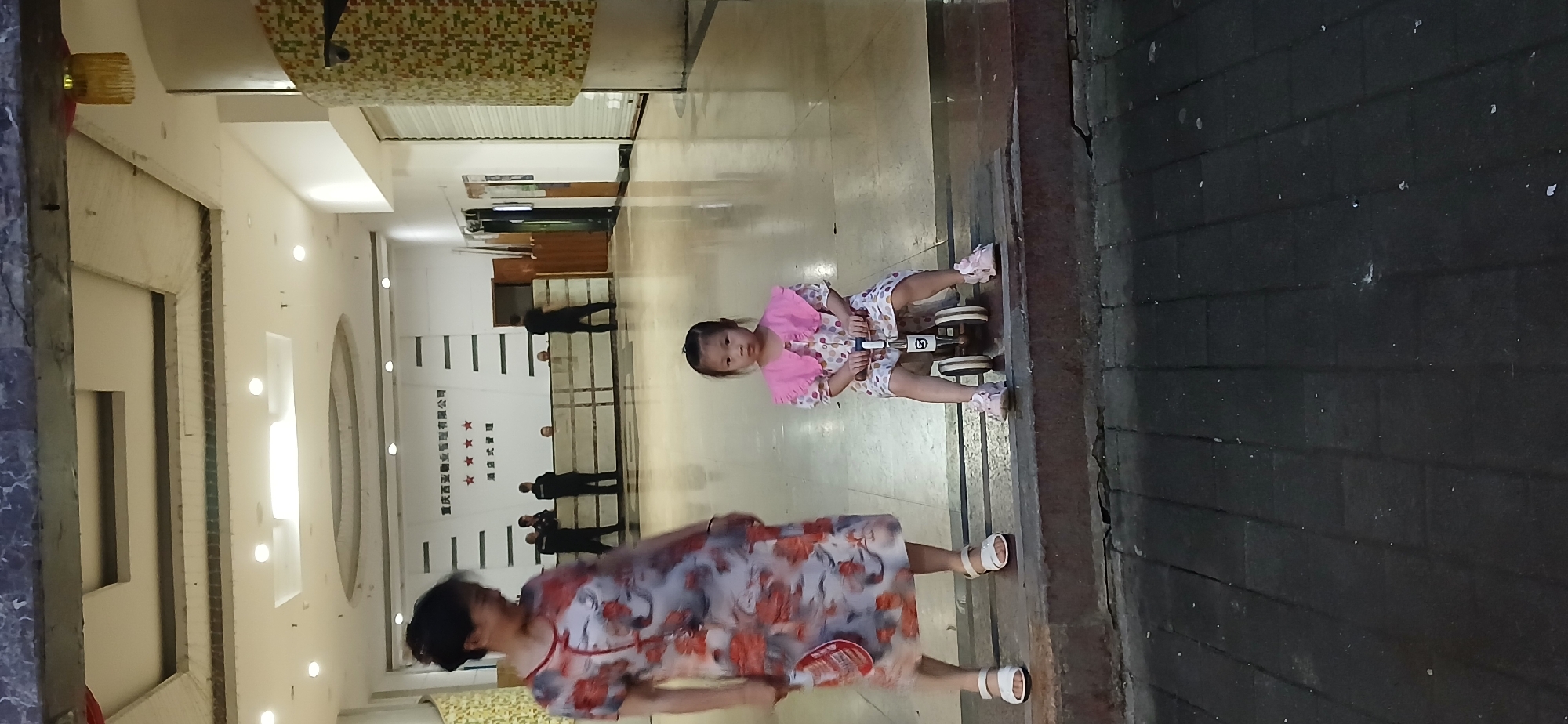 A medium shot shows a woman and a young girl in what appears to be the lobby of a building in Jiulongpo District, China. The woman, on the left, is wearing a floral print dress and white sandals. She is looking to her left and appears to be smiling. The young girl, on the right, is seated on a small tricycle. She is wearing a pink ruffled collar dress with polka dots and pink sneakers. She is looking directly at the camera with a neutral expression. The floor is polished and reflective, indicating an indoor environment. In the background, there is a counter with several individuals standing behind it, some wearing what appear to be uniforms. There are also doors and patterned wall coverings visible. The lighting suggests it might be evening or indoors with artificial light. The overall scene depicts a casual moment with a family member or guardian and a child.
