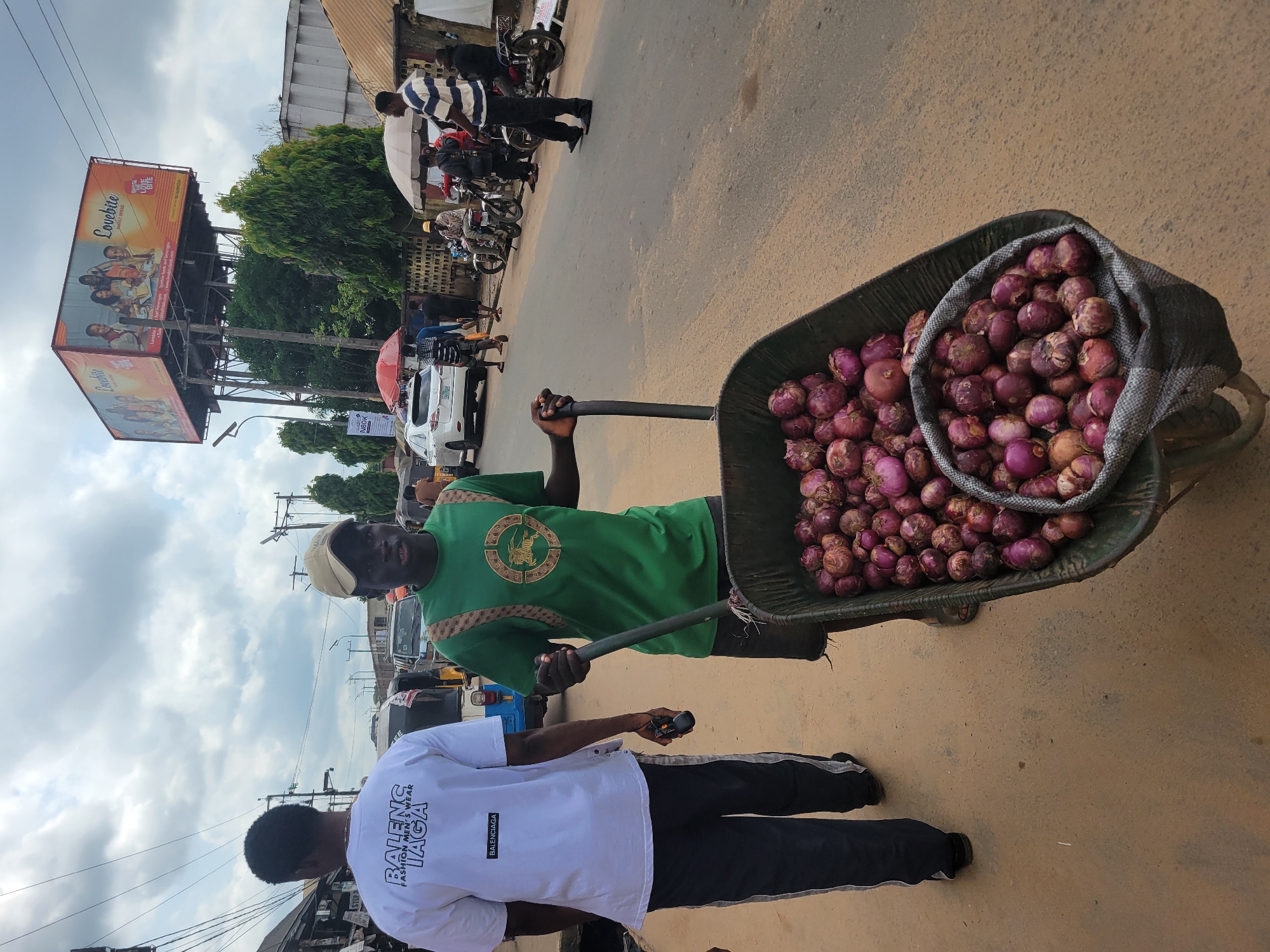 In Egbagu, Nigeria, a man pushes a wheelbarrow overflowing with red onions down a dusty road. He wears a green t-shirt and a beige cap. Beside him, another man in a white t-shirt with "BALENG IAGA FASHION MEN'S WEAR" printed on it walks ahead. The background shows a busy street scene with cars, motorcycles, and pedestrians. A large billboard advertising "Lovebite Family Ahead" is visible in the distance. The sky is cloudy.