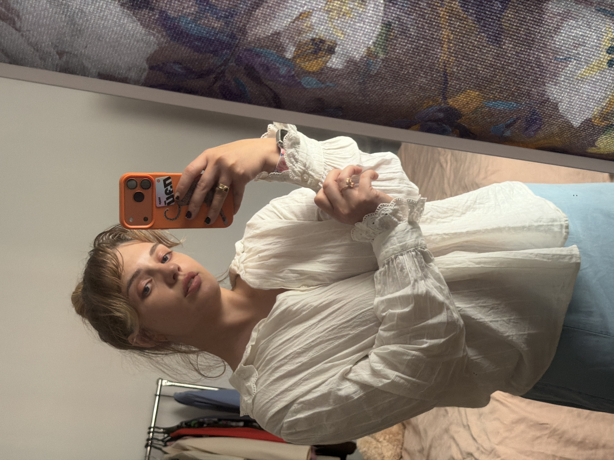 A young woman is captured in a mirror selfie, holding an orange smartphone to take the photo. Her gaze is directed towards the camera, and she wears a neutral expression. She has light brown hair styled with bangs and is dressed in a white, textured blouse featuring ruffled, voluminous sleeves and delicate lace detailing at the cuffs and collar. Rings are visible on her ring fingers, and an Apple Watch with a patterned band peeks out from beneath her left sleeve.
The smartphone case has a sticker on its back that partially displays text, appearing to be "den" or "uen," accompanied by smaller letters "RM" and a decorative swirl. The background of the selfie reflects an indoor setting, likely a private room such as a bedroom or dressing area. To the left, a clothing rack with various garments is visible. Below the woman, light blue fabric, possibly a skirt or pants, is seen, alongside light-colored bedding. Above her head, a blurred reflection of a patterned fabric or object is visible, consistent with a reflection in a mirror. The image was originally rotated 90 degrees clockwise. The setting for this image is Большая Шильна, Russia.