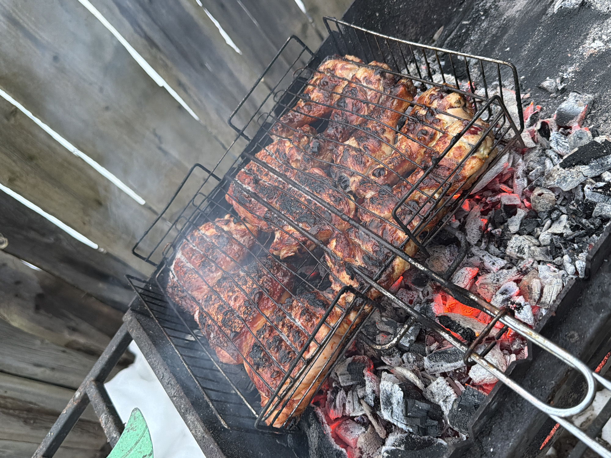 The image shows a metal grill with meat cooking over hot coals. The meat appears to be poultry, possibly chicken, and has grill marks. The grill is positioned above a bed of charcoal, some of which is glowing red. Smoke is rising from the grill and meat. The background includes a wooden fence or wall. It is likely the image was taken outdoors, potentially near the city of Ol'khovo, Russia. No people are visible in the image.