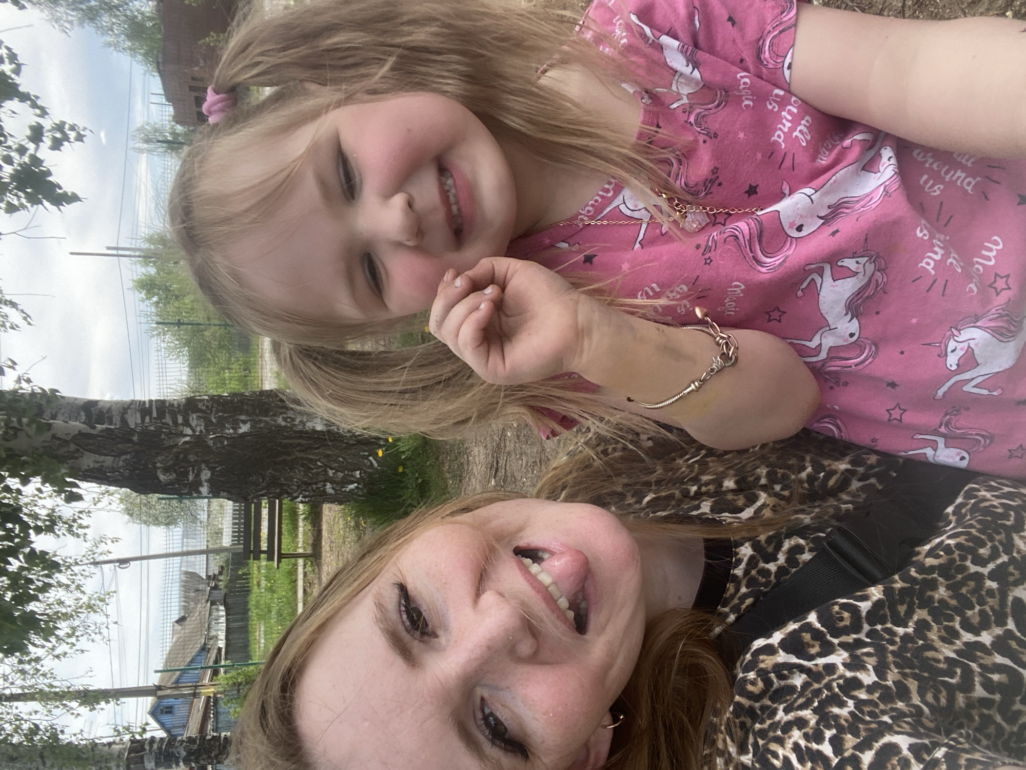 The image is a selfie taken outdoors, likely in a suburban or semi-rural area in the city of Chaikovsky, Russia. A young girl and an adult woman are visible. The girl is wearing a pink shirt with unicorn designs, her blonde hair is in pigtails, and she is smiling. The woman is wearing a leopard print top and sticking out her tongue. In the background are trees, and the corner of a building with a blue roof. A bench and a fence are also visible. Overhead power lines are present, suggesting a residential area.