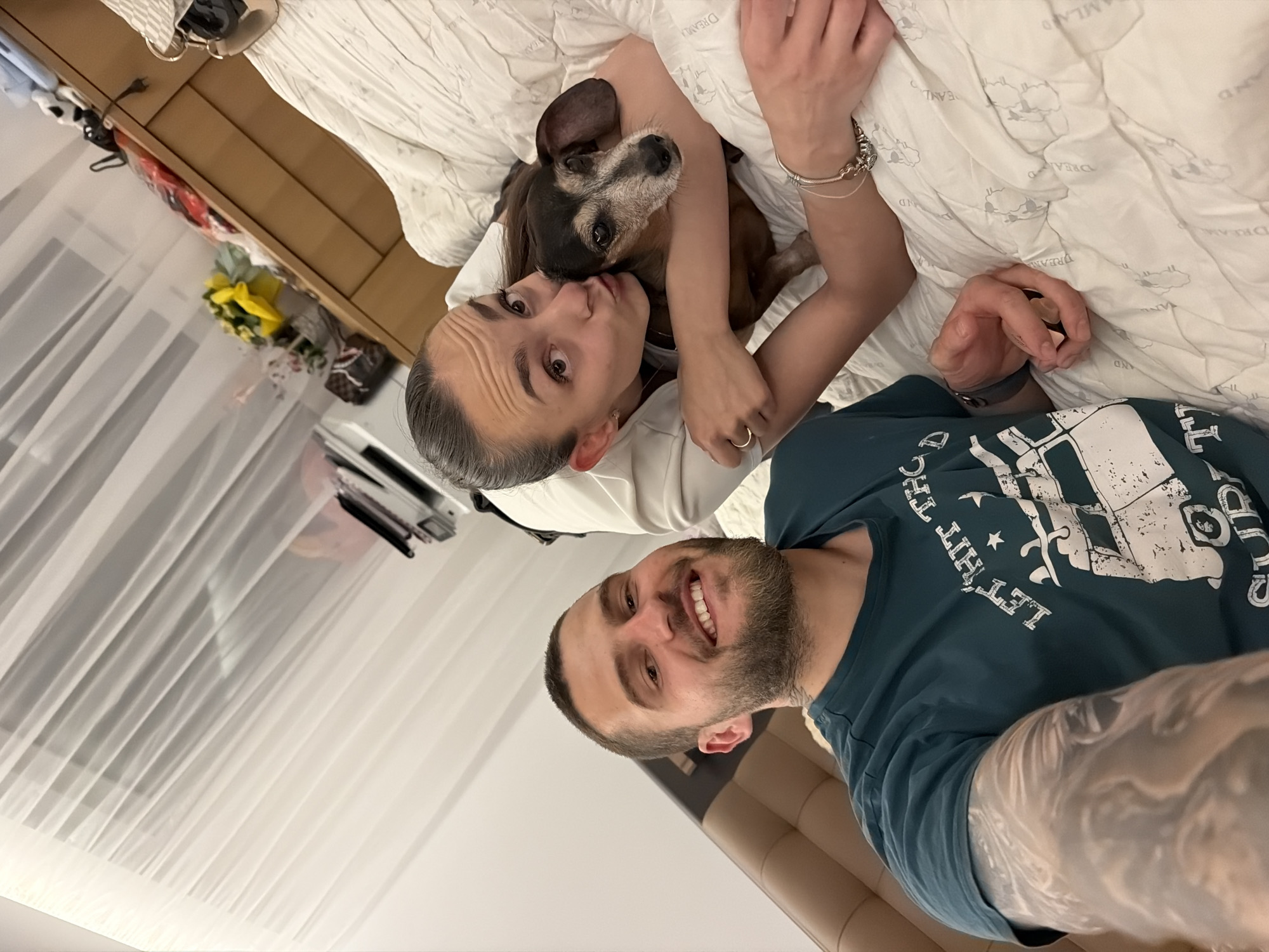 The image depicts a man and a woman on a bed, with a small dog being held by the woman. The man is in the foreground, smiling and looking at the camera. He has short dark hair, a beard, and is wearing a dark teal t-shirt with a graphic design. His left arm, which is tattooed, is visible. The woman is behind him, embracing the dog. She has her hair pulled back and is wearing a white top. She is kissing the dog on the head. The dog is a small dachshund with brown and black fur. The bed is covered with a white, textured comforter. In the background, there is a wooden dresser with various items on top, including flowers and bags, and white curtains on the right. The overall setting appears to be a bedroom. The image does not provide any specific visual cues to indicate the location is the city of Sofia, Bulgaria.