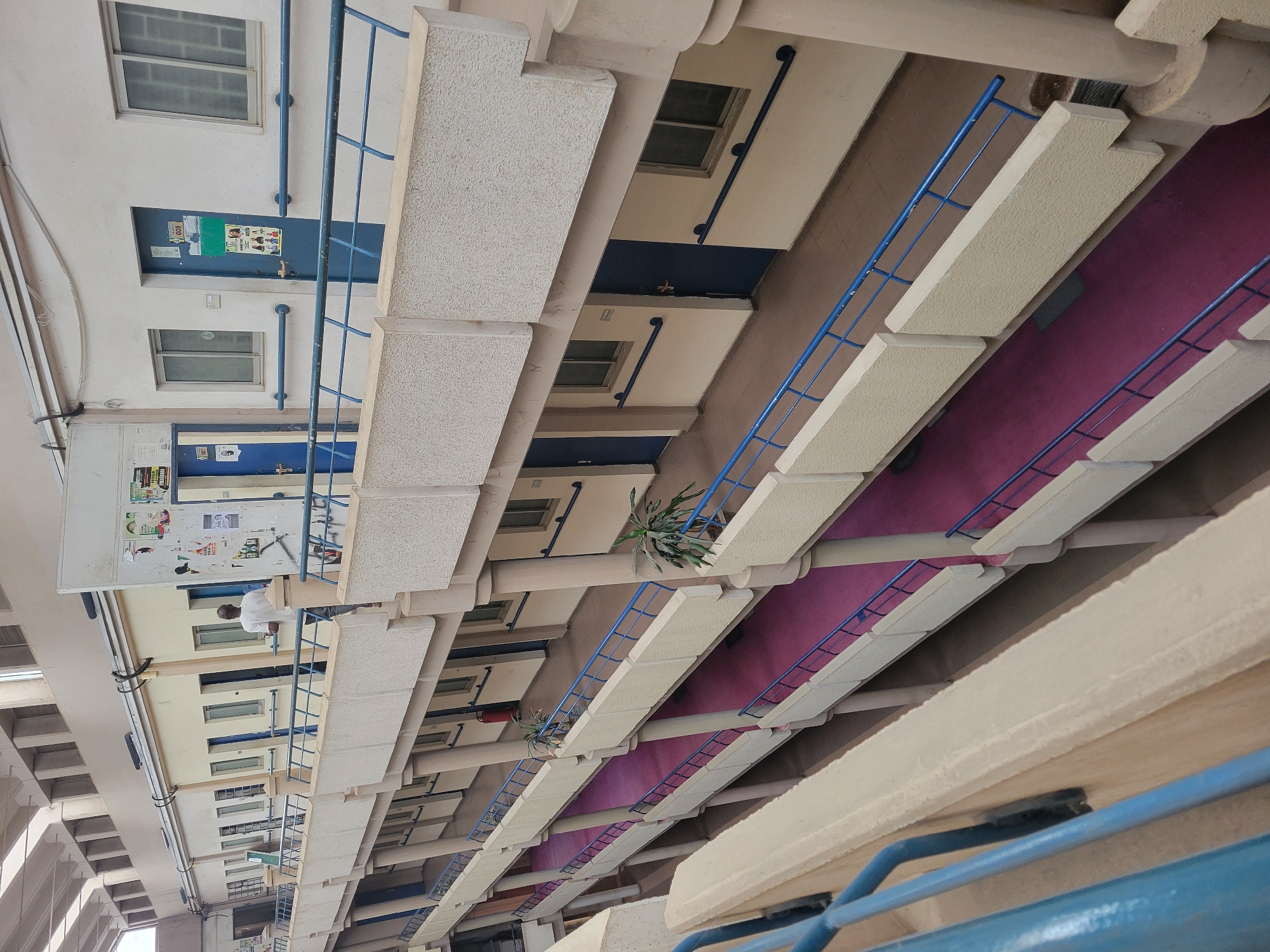The image depicts the multi-story interior of a building, likely a university, office building, or residential complex, in Abuja, Nigeria, viewed from a lower level looking upwards. The structure features several floors, each with a long walkway or balcony lined with blue metal railings. The walkways are distinctly covered in a purple/magenta material.
Along the beige-colored walls of each level, there are rows of blue doors, each flanked by rectangular windows. Whiteboards or notice boards, covered with various papers and posters, are affixed to the walls, particularly prominent on the left side of the frame. One individual, wearing a white shirt, is visible standing on one of the upper walkways.
Decorative green potted plants are placed at intervals along some of the balconies, adding a touch of nature to the environment. A red fire extinguisher is also visible on one of the levels, indicating safety provisions. Utility pipes or conduits run along the walls and ceilings, integrating with the building's architecture. The overall lighting suggests it is daytime, with no direct harsh sunlight, implying an indoor or covered open-air space. While text is present on the notice boards and door signs, much of it is illegible due to distance and resolution, though "ROOM" is partially discernable on one of the blue door signs. The scene portrays an orderly and functional environment.