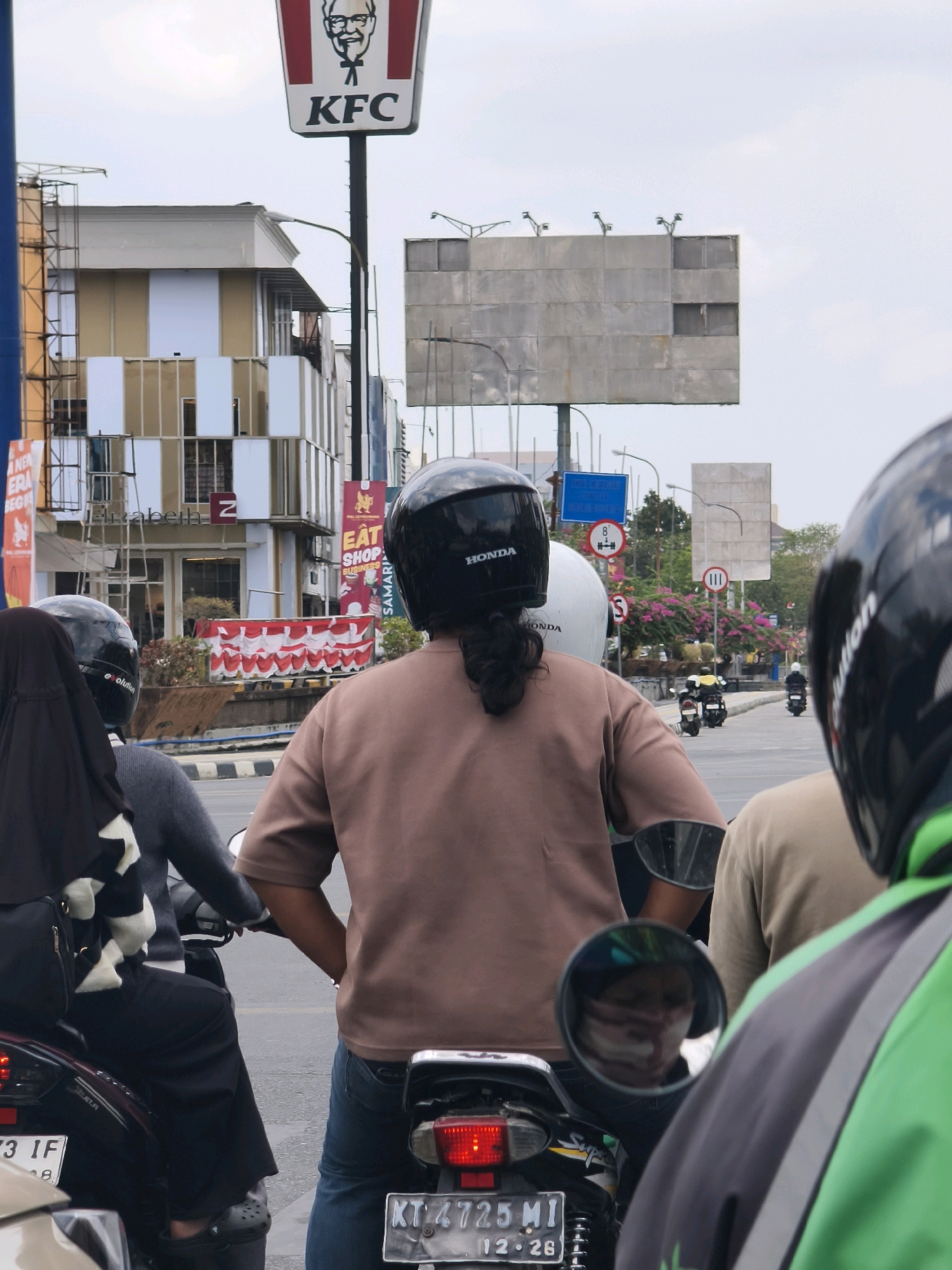 In Samarinda, Indonesia, a group of motorcyclists waits at an intersection. In the foreground, the backs of three individuals wearing helmets are visible. The central figure, wearing a tan shirt and jeans, is looking ahead, while the person to their left, wearing a black hijab and black and white scarf, is also facing forward. A third person in a tan shirt is to the right. Behind them, the red taillights of a motorcycle are visible, along with a license plate reading "KT 4725 MI 12-26".
In the background, a prominent KFC sign stands tall, featuring the iconic Colonel Sanders logo. Further back, a multi-story building with a beige and white facade is visible, adorned with vertical stripes. Various banners and signs are displayed on the buildings, including one with the text "EAT SHOP BUSINESS". Across the street, pink flowers bloom. The sky is overcast. The overall scene depicts a typical urban street in Samarinda.