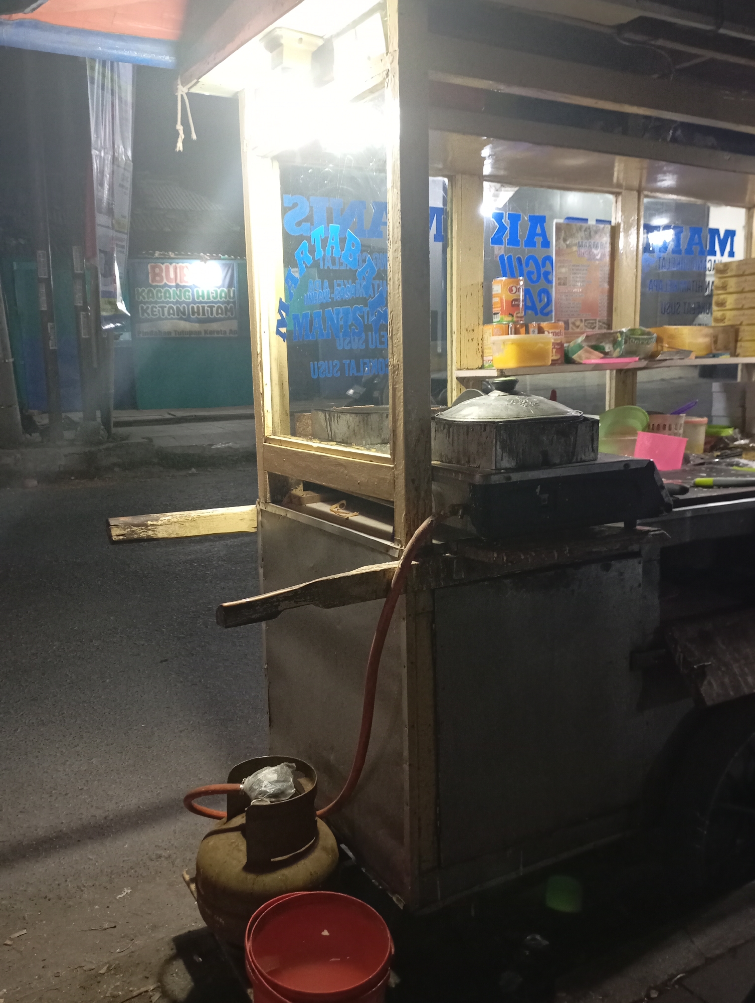 An outdoor, low-angle, night-time photograph shows a food cart at a roadside in Kecamatan Haurgeulis, Indonesia. The cart has a wooden frame with glass panels, illuminating its interior. A large, dark-colored cooking pot sits on top of a scale. A gas cylinder is visible in the foreground, connected to the cart by a red hose. The street is paved and appears wet, reflecting the light. In the background, a sign with text in Indonesian reads "BUBUH KACANG HIJAU KETAN HITAM". Another sign on the cart also has text in Indonesian, partially visible and illuminated, including what appears to be "MANTAP JAYA 2020". The overall scene depicts a small, local business operating at night.