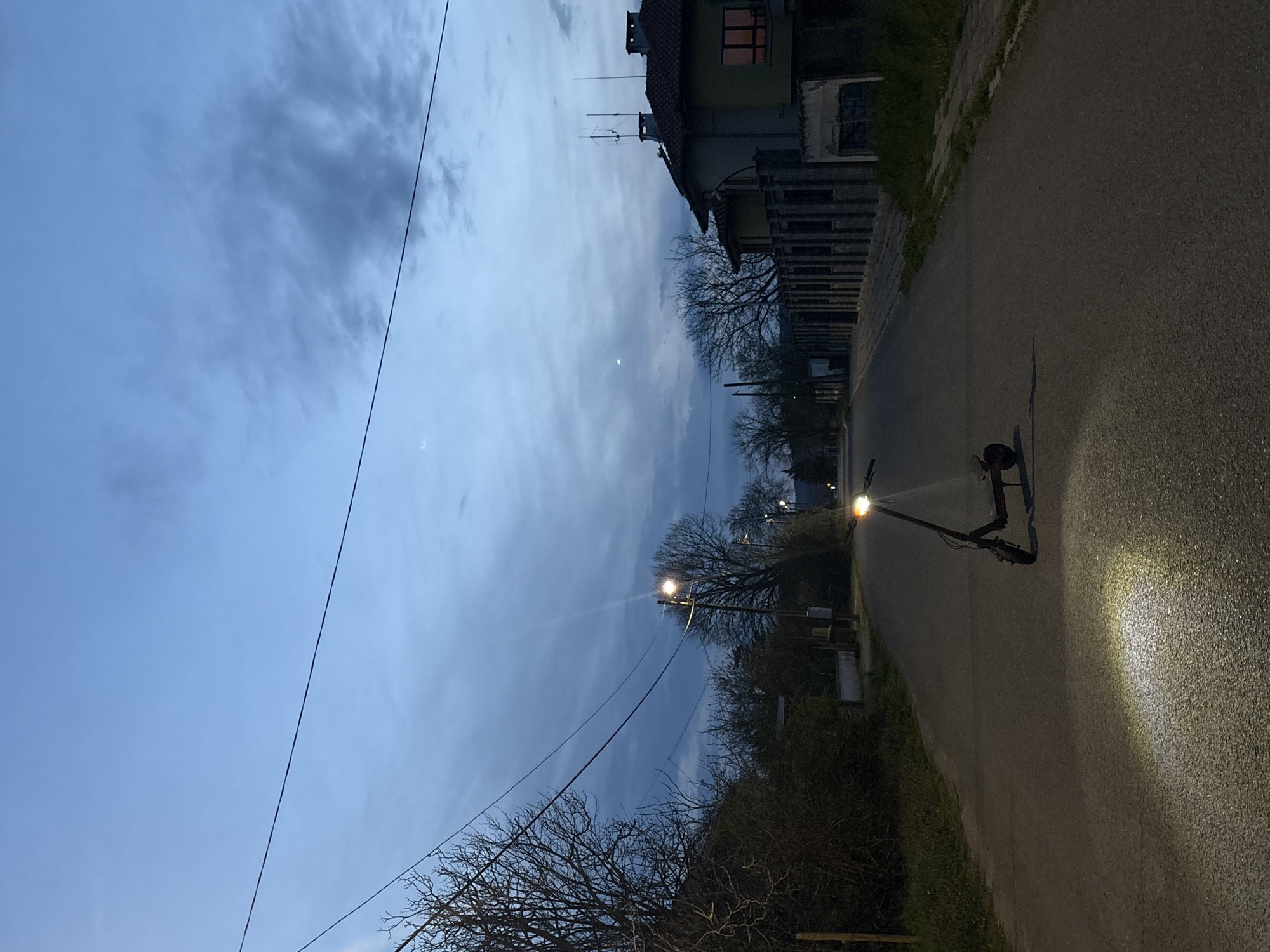 A street scene is captured at dusk or dawn. An electric scooter with its headlight illuminated is parked on the right side of a paved road. The road runs between buildings on the left and foliage on the right. Utility wires stretch across the upper left of the frame, cutting through the sky. The sky is overcast with clouds, and a faint crescent moon is visible. Streetlights are on, casting a glow amidst the trees.