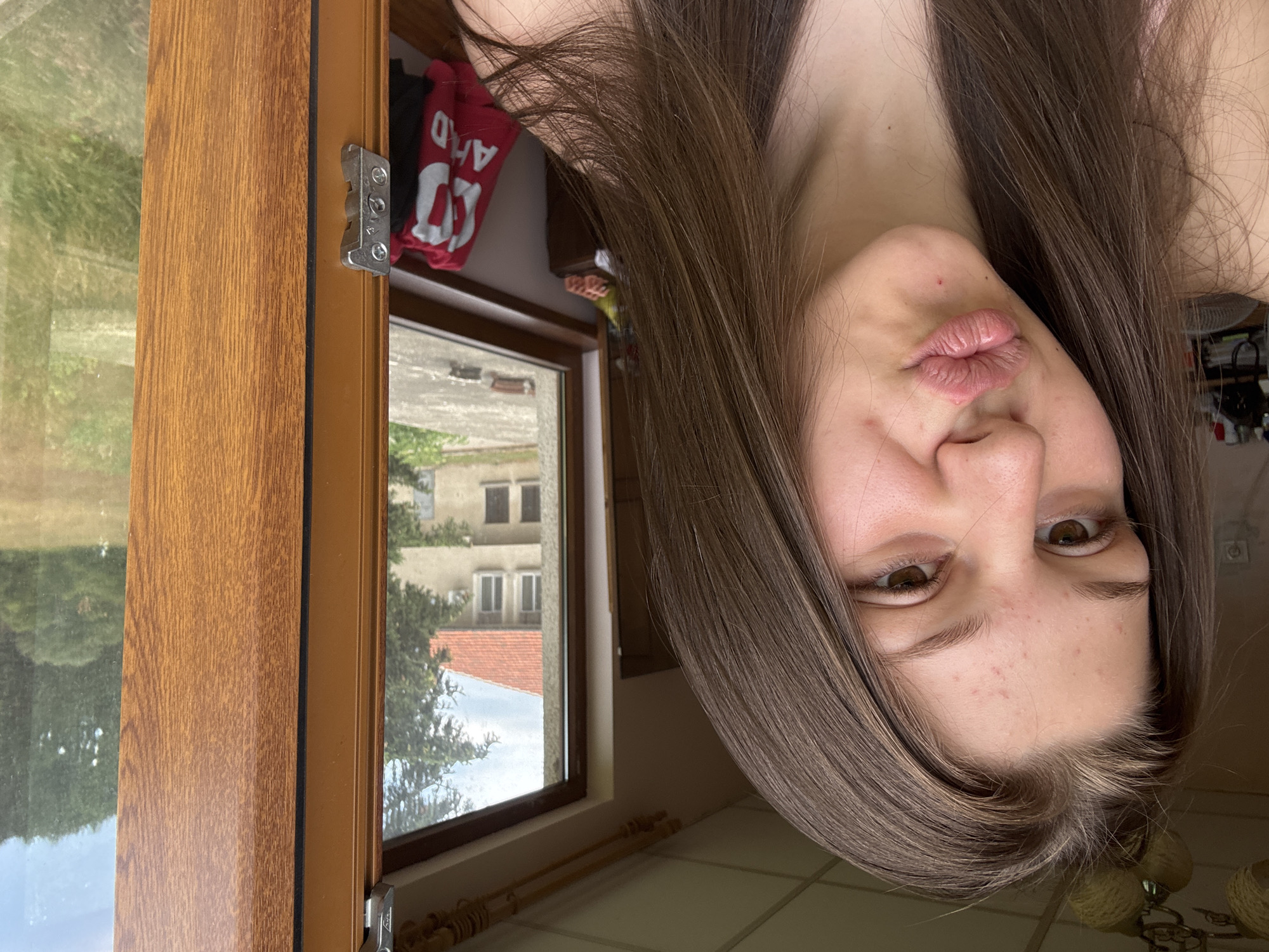 A young woman with long brown hair is captured in a close-up selfie, puckering her lips. She is positioned on the left side of the frame, with her face and upper body visible. The background reveals a window with a view of a residential building with a red-tiled roof and a lush green landscape. The window frame is made of wood, and a metal latch is visible on the left side. Inside the room, a red garment with white lettering is draped over a piece of furniture. The lighting suggests an indoor setting with natural light coming from the window.