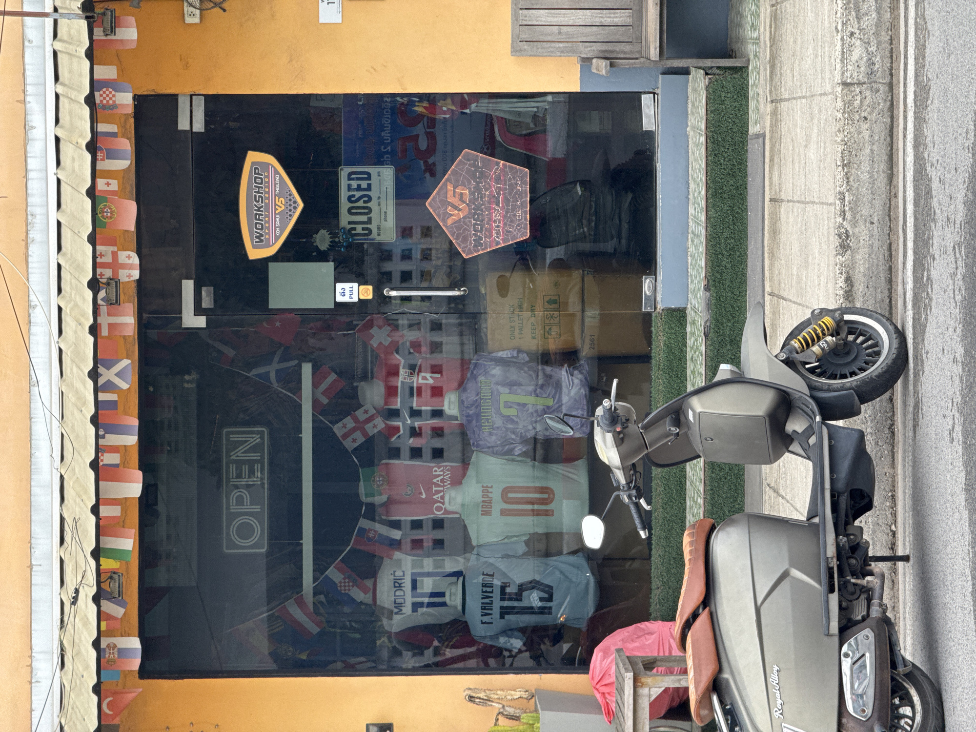 The image shows the exterior of a shop with a large glass storefront. A scooter is parked on the sidewalk in front of the shop. The shop displays several sports jerseys on mannequins inside. A string of international flags hangs above the storefront. A "CLOSED" sign is visible on the glass door, along with a "PULL" sign. The building's exterior is painted orange. A wooden bench is situated to the right of the shop entrance.