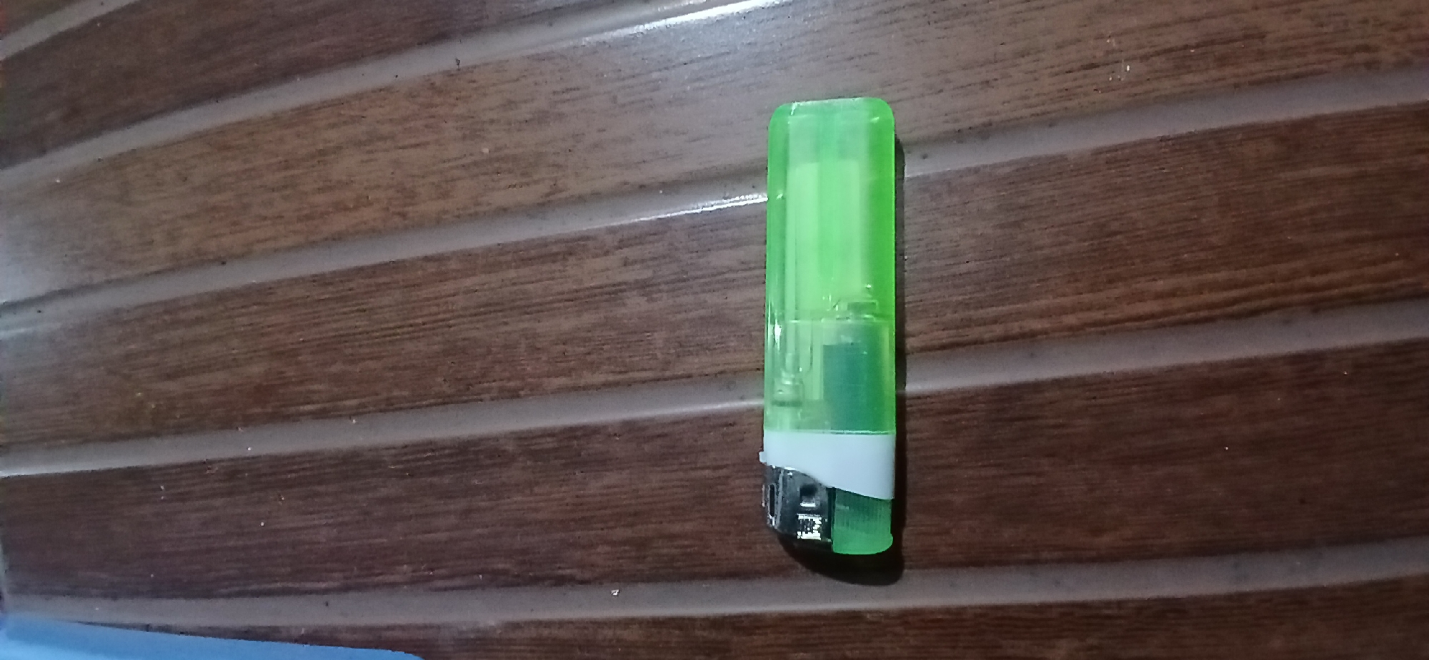 A green and clear plastic lighter lies horizontally on a wooden surface with a wood grain pattern. The lighter has a metallic top and a translucent green body. The surface appears to be a table or countertop. The lighting is dim, casting shadows on the surface. There are no people or other objects visible in the image. The location is Sekarputih, Indonesia.