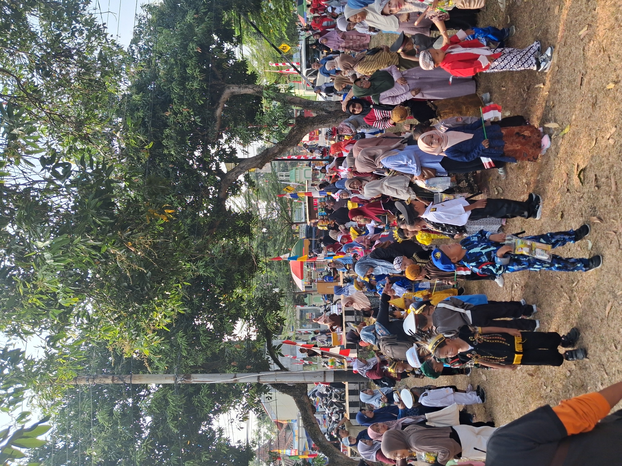 The image depicts a crowded outdoor event in Balapulang, Indonesia, likely a local celebration or festival. The scene is filled with people, predominantly women and children, many of whom are wearing traditional Indonesian attire, including headscarves and batik patterns. Several children are dressed in costumes resembling military or traditional uniforms, some holding small Indonesian flags.
The environment features large trees providing shade, with evidence of electrical wires and poles overhead. In the background, there are buildings, parked motorcycles, and colorful decorations including flags and umbrellas. The ground is covered in dry leaves and dirt, suggesting a park or an open public space. The overall atmosphere appears festive and communal, with people gathered in groups, possibly for a parade or a performance. The presence of Indonesian flags and the traditional clothing strongly indicates a local cultural event.