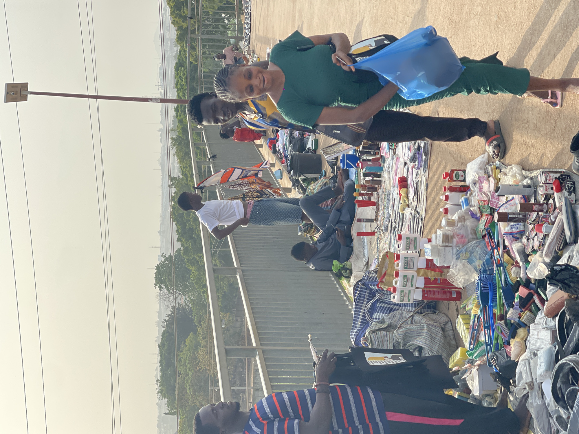 On an elevated concrete surface, likely a pedestrian walkway or bridge, a bustling open-air market is teeming with vendors and customers. The location is Kuchingoro, Nigeria, as implied by the prompt. The market displays a variety of goods spread out on cloths or mats.
At the forefront, a woman in a green top is holding a blue plastic bag, facing the camera with a smile. Behind her stands a man in a yellow and blue shirt. Several other individuals are visible amongst the market stalls, including a man in a white shirt. One man is seated while browsing through the items, another man stands wearing a blue and red striped shirt.
The goods on display include items packaged in red and white containers, possibly health products or household supplies. Also visible are brushes and assorted cleaning supplies. The background reveals a hazy cityscape and trees. Power lines are stretched across the sky, adding to the urban setting. A vertical post is visible on the left of the image.