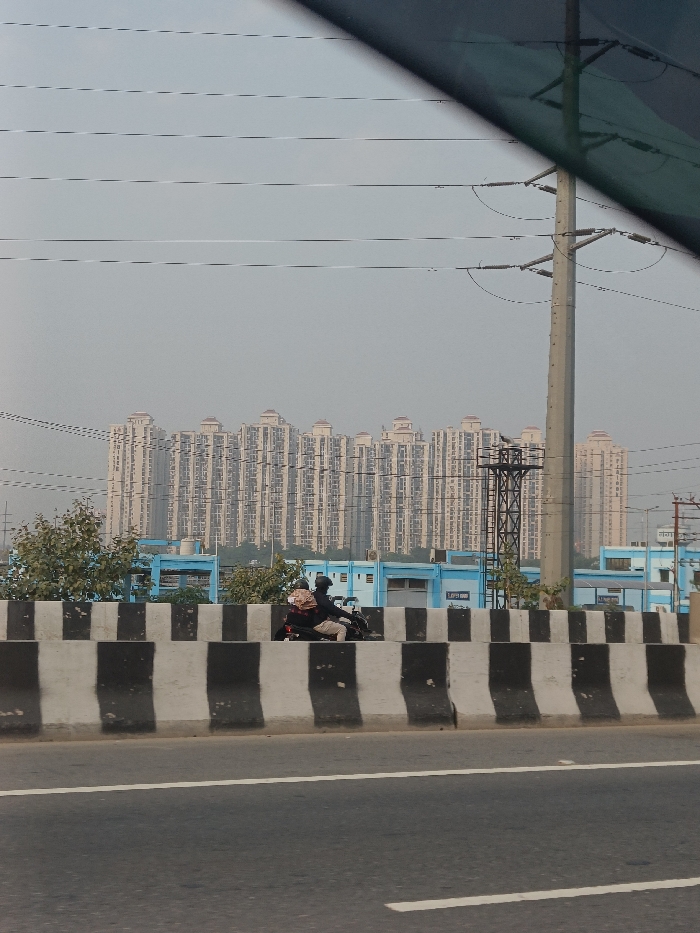 This image captures an urban landscape in Ghaziabad, India, featuring a blend of infrastructure, residential buildings, and daily activity under a hazy sky. The photograph appears to be taken from a moving vehicle, with a dark section of a car window or roof visible in the upper right corner.
In the immediate foreground, a wide asphalt road is visible. A prominent concrete barrier with alternating black and white stripes runs across the midground, separating the main road from a lower-level path or service road. On this path, two individuals are seen riding a motorcycle. The driver wears a dark helmet and jacket, and the passenger, dressed in lighter attire, has a brown backpack or parcel. Sparse green foliage and bushes are visible behind the barrier to the left.
Behind the barrier, a series of low-rise buildings with blue exterior walls and white roofs are present. Text, possibly reading "LAPP OMN," can be faintly discerned on one of these blue structures. To the right, a tall, grey utility pole stands, supporting multiple power lines that extend horizontally across the frame.
Dominating the background is a long row of numerous tall, light-colored residential apartment buildings. These multi-story structures exhibit similar architectural styles with many windows and balconies, forming a significant urban development against the horizon.
The sky is a uniform, pale grey, suggesting an overcast day or potentially hazy/smoggy atmospheric conditions, common in the region. The scene depicts a typical daytime urban environment with ongoing movement and extensive building development.