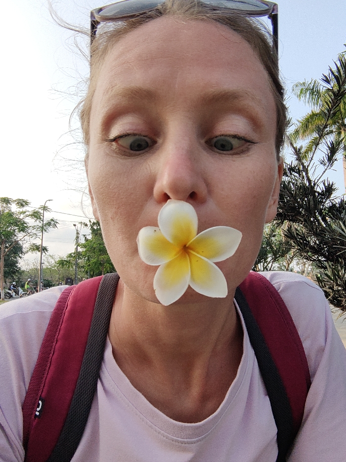 A close-up portrait shot in Thuận Hóa Ward, Vietnam, features an adult female subject from the chest upwards. She has fair skin and light-colored hair, with dark sunglasses resting on top of her head. Her expression is characterized by wide-open eyes looking downwards, and a white Plumeria flower with a yellow center is positioned between her nose and upper lip, covering her mouth. She wears a light purple crew-neck t-shirt and a maroon backpack with a dark grey padded strap, visible over her left shoulder. The outdoor background on the left includes deciduous trees, a street light pole, and several distant figures on motorbikes on what appears to be a road under a bright, overcast sky. On the right, a large palm tree and dense dark green foliage are visible.