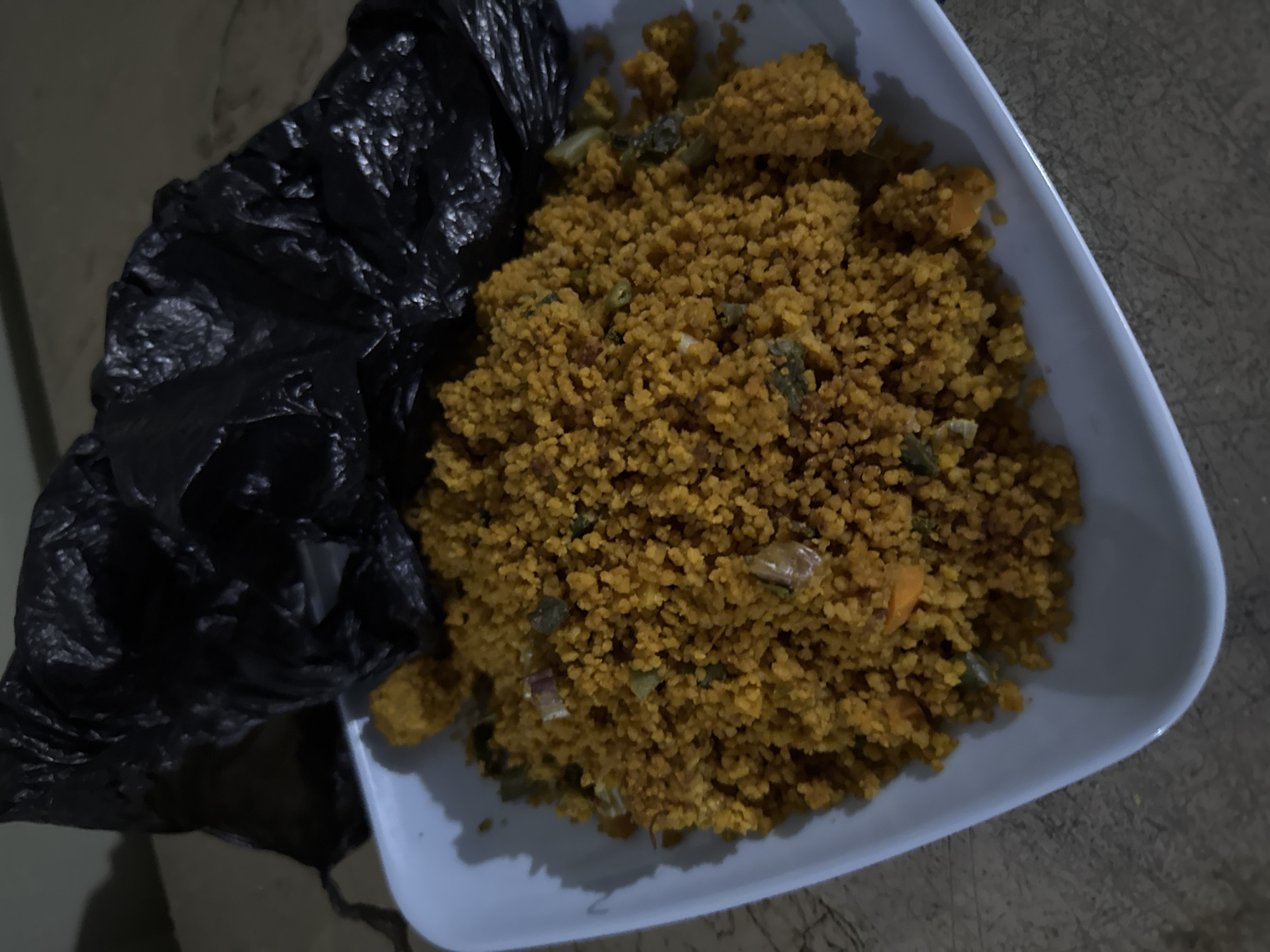 An overhead shot captures a light blue platter filled with a mound of yellow couscous mixed with chopped vegetables. The couscous has a granular texture, and visible within it are pieces of what appear to be carrots, green herbs, and possibly onions or peppers. To the upper right of the platter, a crumpled black plastic bag is positioned, its dark form contrasting with the lighter colors of the food and dish. The platter rests on a textured surface, possibly a countertop or table, with a muted brown and beige color scheme. The lighting appears dim, casting shadows and highlighting the textures of the food and the plastic bag.
