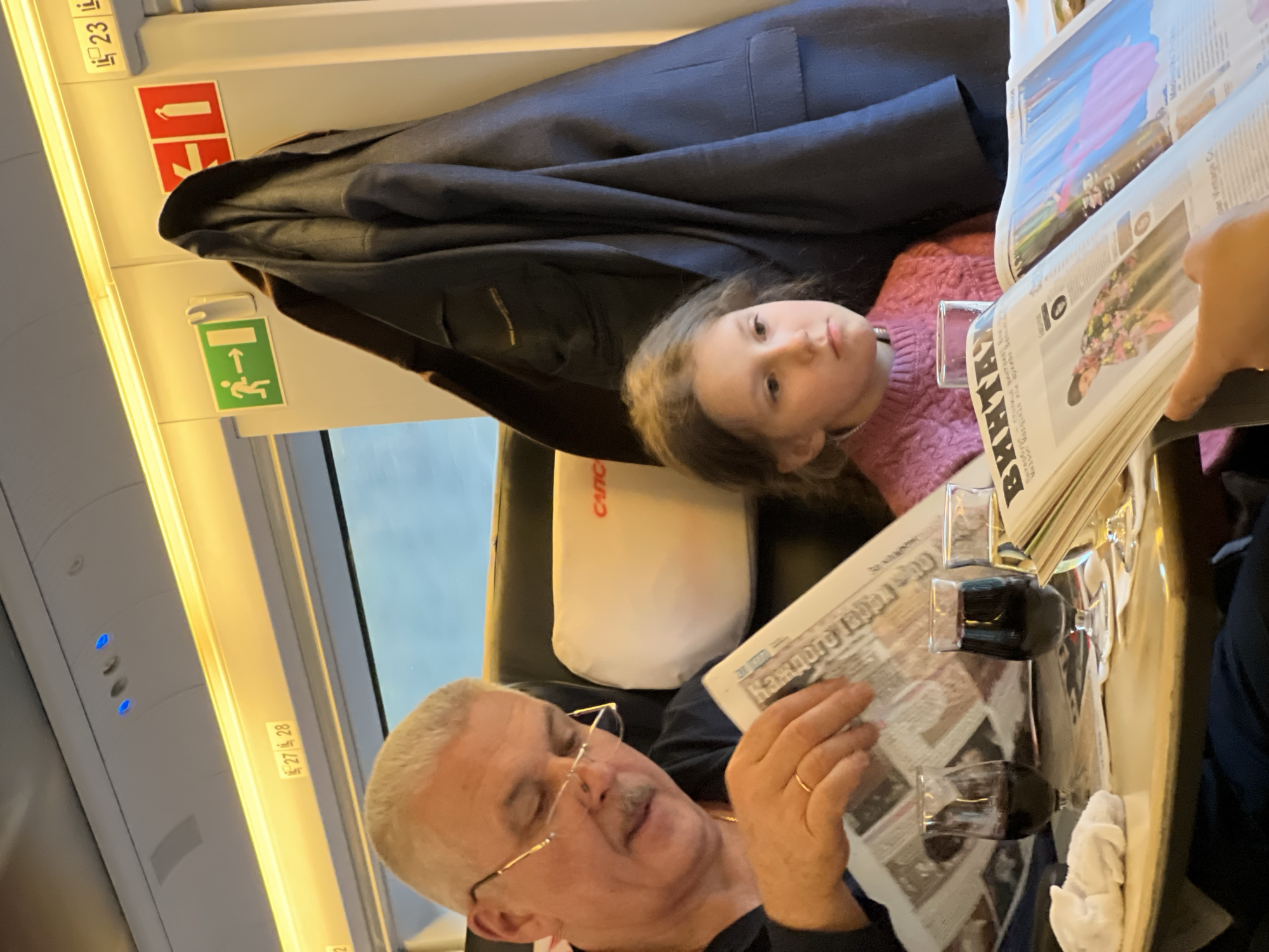 On a train, a man in glasses reads a newspaper, with a young girl next to him. A dark jacket hangs above them. Two glasses of dark liquid are on the tray table. The train interior includes overhead lights and emergency signs. It is not possible to determine if this is Reshetnikovo, Russia, without additional information.