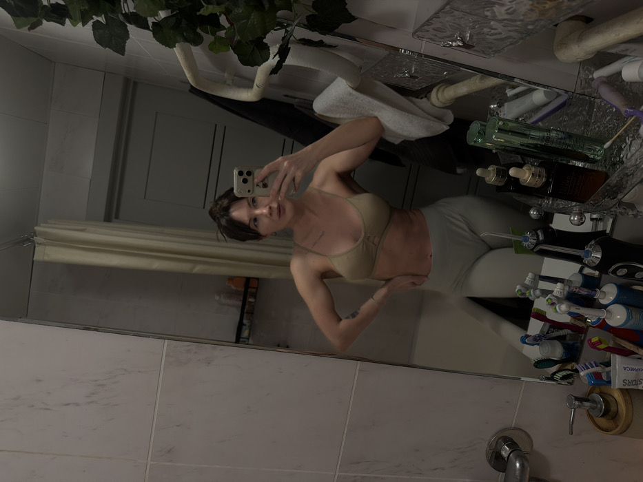 A female subject is captured in a mirror selfie taken within a residential bathroom. The subject, appearing to be an adult with dark hair styled in a bun, is wearing a light tan bralette and light grey leggings. Her left hand rests on her hip, while her right hand holds a light-colored smartphone, positioned to photograph her reflection. Tattoos are visible on her left bicep and potentially on her upper chest.
The bathroom features light-colored wall tiles, possibly white or cream with subtle grey veining. A bathtub is partially visible in the background, obscured by a light-colored shower curtain. The mirror reflects a portion of the room, including a door with two recessed panels. To the right of the subject, a white, serpentine heated towel rail is affixed to the wall, draped with a white towel and partially covered by an artificial green leafy plant.
The counter below the mirror is cluttered with various toiletries and personal hygiene items. These include multiple toothbrushes (both manual and electric), a water flosser, a tube of toothpaste with Cyrillic text ("SPLAT" or similar), a pump-style soap dispenser with a wooden base, and several bottles of cosmetic products, notably a brown dropper bottle resembling "Estée Lauder" and a tall green bottle. The presence of Cyrillic text on the toothpaste tube indicates the location is consistent with Russia. However, specific visual identifiers to confirm the city of Perm within Russia are not present in the image.