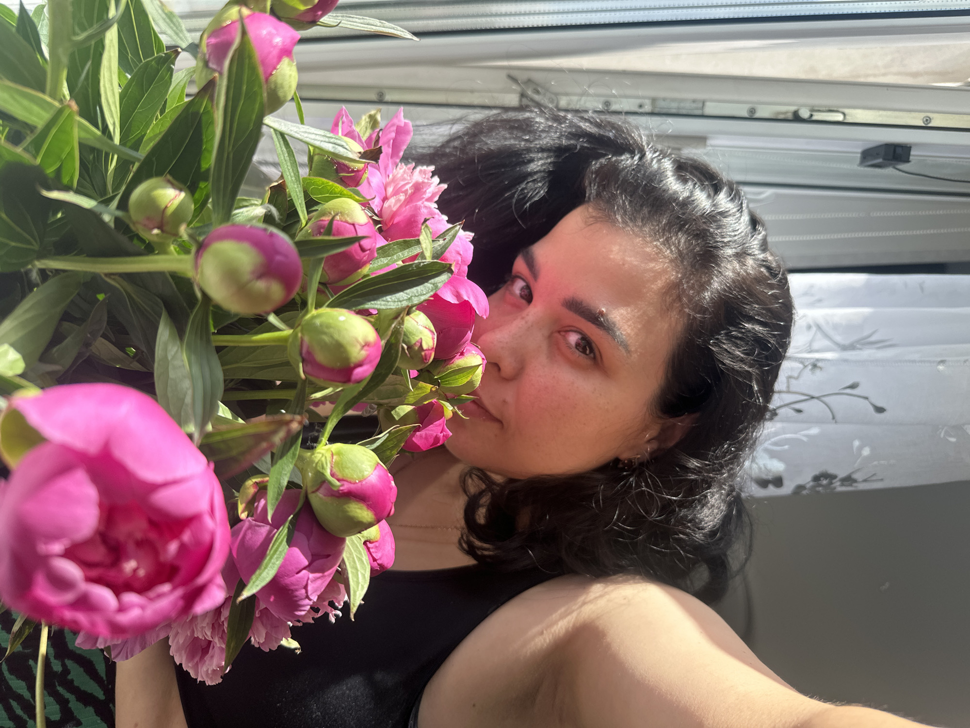 A close-up, top-down perspective depicts one individual, appearing female, positioned in the foreground peering over a bouquet of vibrant fuchsia-pink peonies. The individual has dark, curly hair, dark eyes looking directly into the camera, and a fair complexion with visible freckles. They are wearing a black sleeveless top, with a portion of a green and black patterned garment visible at the bottom right. The bouquet consists of multiple peonies, with several still in bud form (green with pink tips) and others partially or fully open, displaying their ruffled petals. Green leaves are prominent within the bouquet. The background features an indoor setting illuminated by strong natural sunlight. To the left, a white window frame with vertical blinds and a small black electronic device attached near the top are visible. To the right, a sheer white curtain with a subtle dark botanical pattern covers another window area, behind which a darker, plain wall is present. The lighting casts highlights on the person's skin and hair. The location is in the city of Unknown, Unknown.