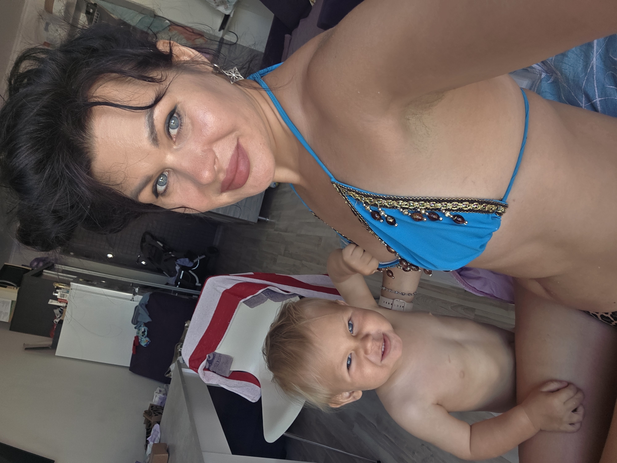 In this indoor, low-light, portrait-oriented selfie, a woman with dark hair and blue eyes, wearing a blue bikini, holds a young child. The woman's hair is dark and appears styled in waves. She is looking towards the camera with a slight smile, her makeup visible. The child, with blond hair and blue eyes, is also looking towards the camera and smiling broadly. The background shows an interior scene with what appears to be a floor and furniture. There is a towel with red and white stripes draped over a white object in the lower left portion of the frame. The scene suggests a casual, possibly domestic setting. There is no visible text.