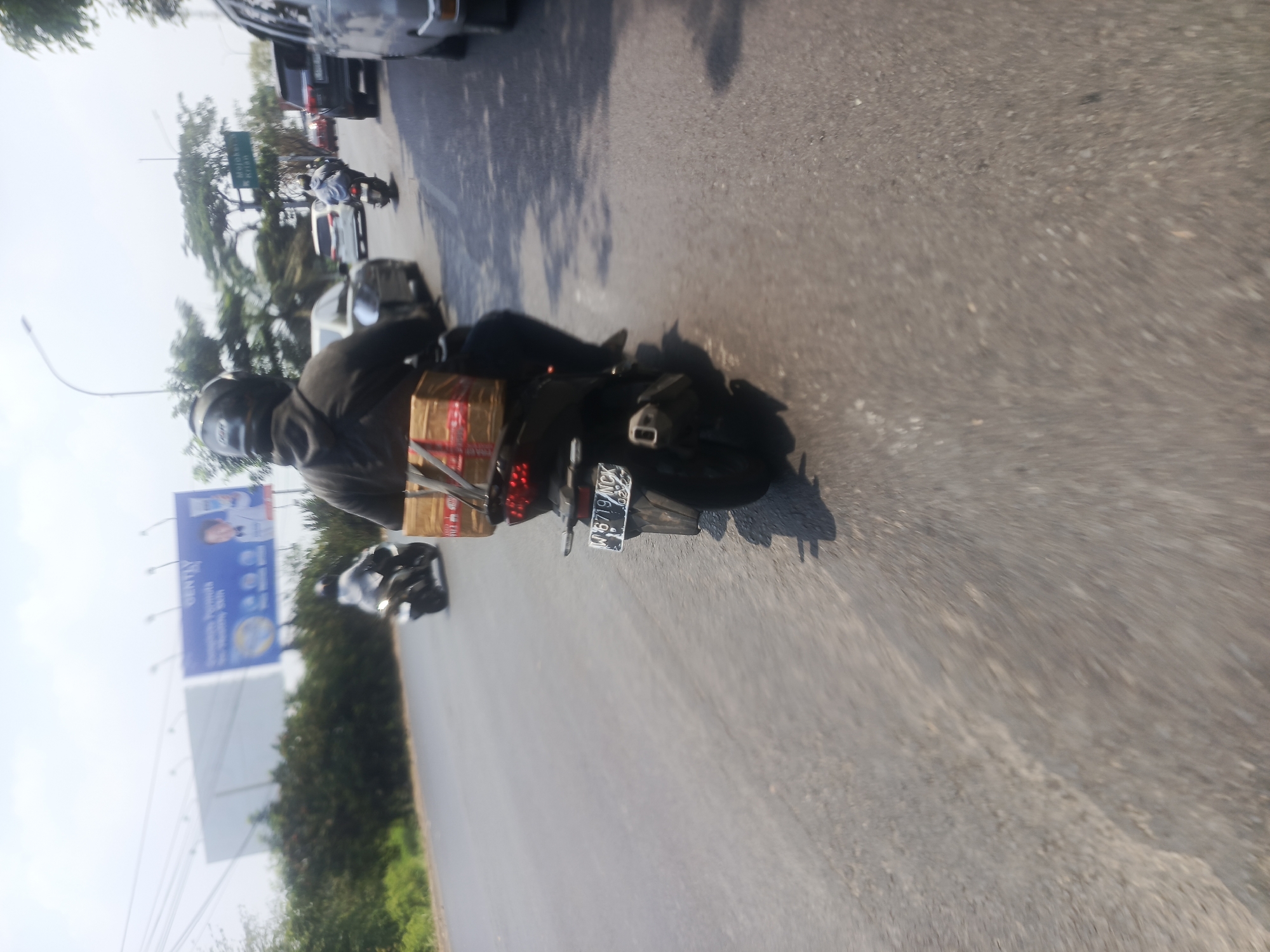 The image depicts a street scene in Surabaya, Indonesia, captured from the perspective of someone riding a motorcycle. The foreground shows the back of a motorcycle with packages strapped to it and the rider wearing a black helmet and jacket. A license plate with the characters "W 6719 NCX" is visible on the motorcycle. In the background, other vehicles, including cars and motorcycles, are present on the road. A billboard advertising "GENTLY" and a portrait of a woman is visible on the left side of the image. Trees and buildings line the street, with a bright sky overhead. The scene appears to be captured during daylight.