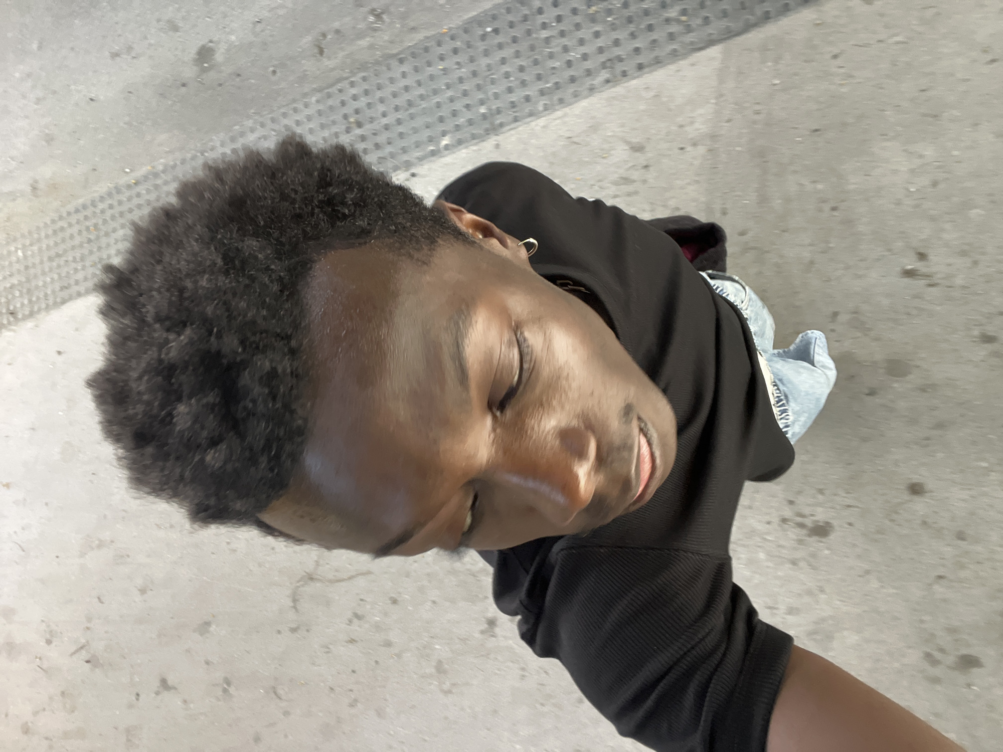 This media file features a close-up, overhead shot of a young Black person. They are wearing a black hooded sweatshirt and light-wash denim jeans. The person has short, dark, curly hair and is wearing a hoop earring in their visible ear. Their eyes are closed, and they have a slight smile on their face, suggesting a relaxed or content emotion. The lighting indicates it is daytime, and the scene appears to be outdoors on a concrete surface. In the background, there is a textured strip of gray material with a pattern of small circles, possibly a grate or textured paving. The ground surface is speckled with dirt and debris. The overall setting is urban and appears to be a public space, with the location context suggesting Sant Adrià de Besòs, Spain. No visible text is present in the image.
