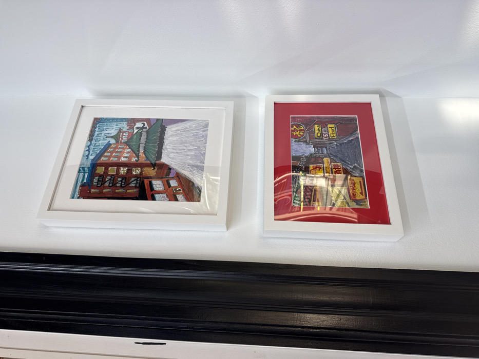 Two framed artworks are vertically mounted on a white wall. To the left, a black architectural trim is partially visible. The upper artwork, a portrait-oriented piece in a white frame with white matting, depicts a multi-story red-brick building adjacent to a green-tiered pagoda-style roof, indicative of the Chinatown Gate. The lower artwork, a landscape-oriented piece in a white frame with red matting, illustrates a narrow street scene with buildings and multiple illuminated signs featuring Asian characters and English text, including "Chinatown" and "China Pearl." Both artworks distinctly portray elements of Chinatown in Boston, Massachusetts, United States. Reflections are visible on the glass of the lower frame, suggesting ambient light. No people are present, and no actions or interactions are occurring within the visible scene.
