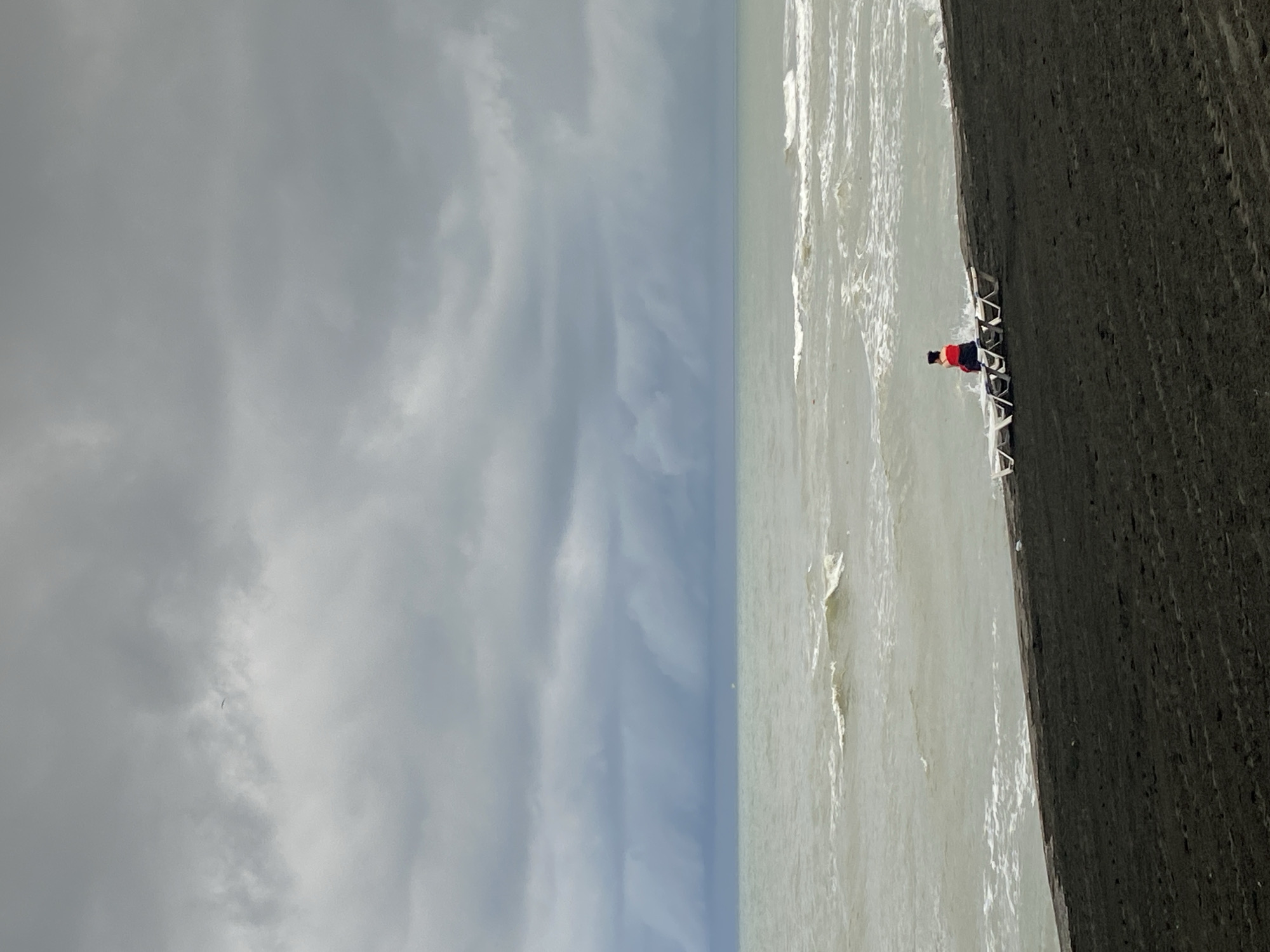 The image depicts a solitary person sitting on a beach chair on the black sand beach of Ureki, Georgia. The person is wearing a red top and dark bottoms. Several other empty beach chairs are arranged in front of them. The sea is visible with choppy waves, and the sky is overcast with heavy, dark clouds. The overall atmosphere suggests a calm, possibly impending weather change.