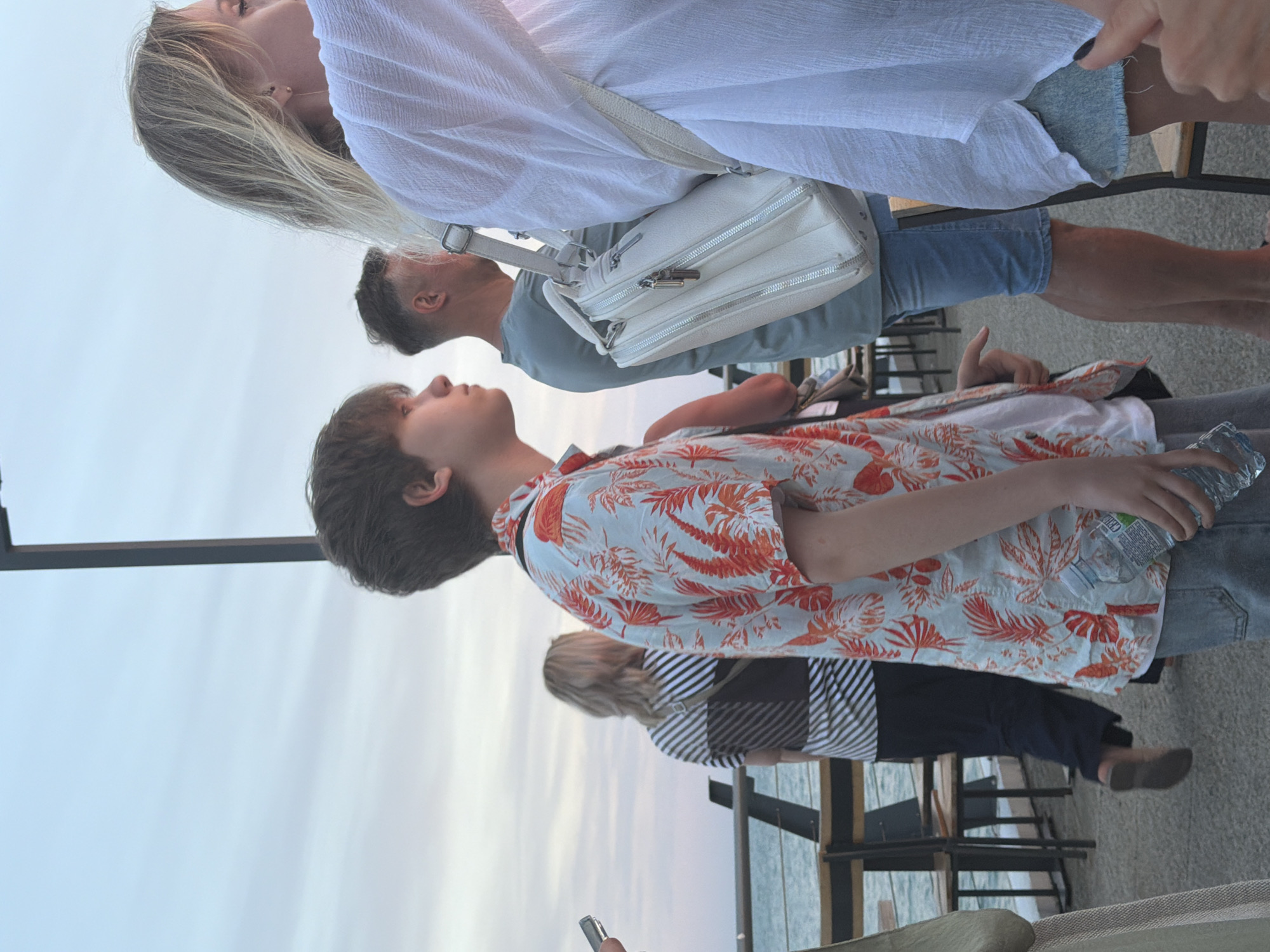 The image shows four people standing outdoors. The primary subject is a young person wearing a short-sleeved, brightly patterned shirt with red and white foliage designs. They are holding a clear plastic bottle of water in their right hand. To their left, a person with short, dark hair is visible, looking upwards and to the left. Behind them, a woman with long blonde hair is also looking upwards, and she has a white purse slung over her shoulder. The fourth person is partially visible behind the woman, wearing a striped shirt. The setting appears to be a waterfront or outdoor dining area, with tables and chairs in the background, suggesting a casual gathering or meal. The sky is a pale blue with hints of pink, indicating it might be late afternoon or early evening. The weather seems pleasant. The overall mood is relaxed and casual. No discernible text is visible in the image.