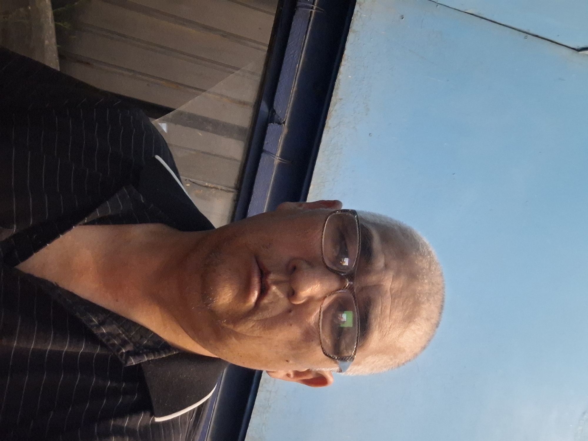The media file contains a portrait of an older Caucasian man with fair skin and grey hair, wearing glasses and a dark pinstriped shirt. He is looking directly at the camera, with a neutral expression. The man is positioned in the foreground, with his face and upper body visible. The background is out of focus and appears to be an outdoor setting with a light blue sky and some horizontal wooden structures. The lighting suggests it might be daytime, possibly late afternoon due to the warm tone of the light on the man's skin. No text is visible in the media file. The image is oriented vertically.