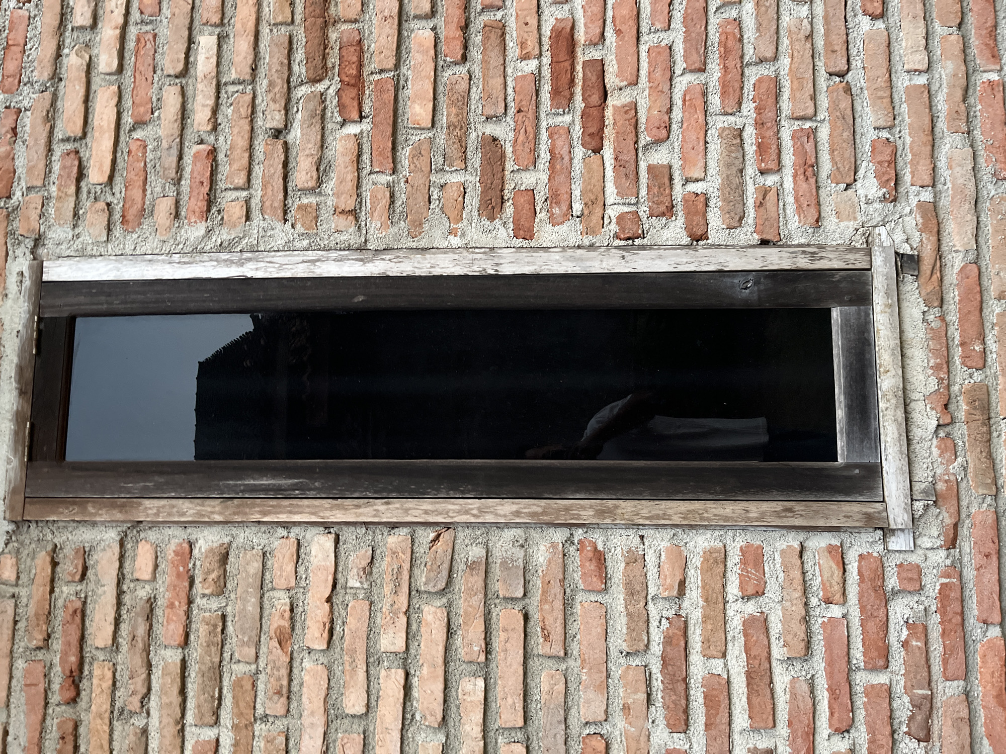 This image shows a tall, narrow window with dark glass set into a brick wall. The bricks are reddish-orange and appear to be old, with some variations in color and texture. The mortar between the bricks is gray and rough. The window frame is made of wood, which is weathered and appears to be dark brown. A reflection of a person wearing a light-colored shirt can be seen in the glass. The scene suggests an outdoor setting, possibly a building in Kebumen, Indonesia, given the context.