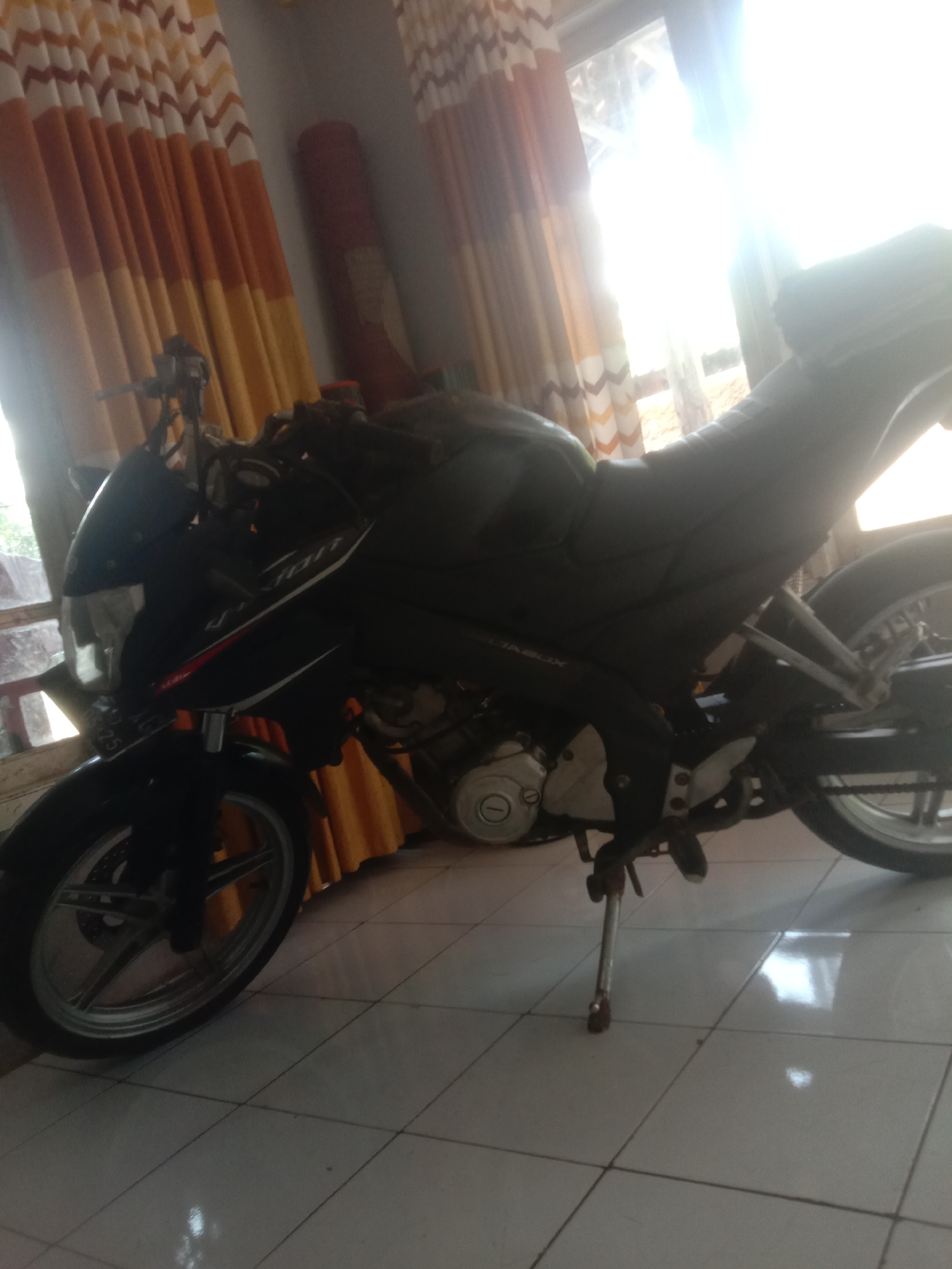 A dark-colored motorcycle is parked indoors, with its front wheel facing left and its rear wheel facing right. The motorcycle is angled slightly towards the camera. The handlebars are visible, as well as part of the seat and the engine. The motorcycle has a license plate on the front. The background consists of a tiled floor and orange and yellow curtains hanging on the right side of the image. A large window with bright sunlight shining through is to the right of the motorcycle. The lighting in the image is uneven, with strong highlights and shadows. The overall scene appears to be an interior shot within a residential or commercial space in Cianjur, Indonesia.