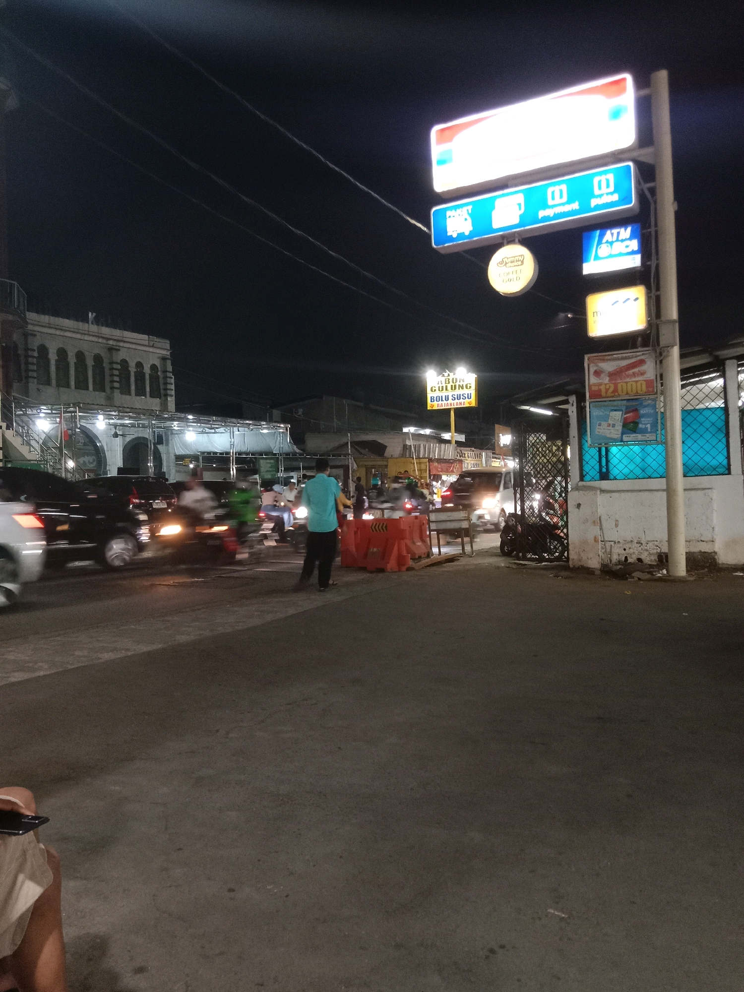 A night scene depicts a busy street in South Jakarta, Indonesia, likely near a minimarket or convenience store. Several signs are illuminated, including one that reads "ATM BCA" and another with "payment pulse." There are also signs for "PAKET" (package) and "GULUNG BOLU SUSU RAIAKANA" (rolled milk cake).
Vehicles, including cars and motorcycles, are visible on the road, some of which are in motion, causing motion blur. Several individuals are present. One person in a teal shirt stands near orange traffic barriers, appearing to direct traffic or manage the area. Another person is seated in the foreground on the left, holding a smartphone. The overall environment suggests a commercial area with ongoing activity. The architecture of a building in the background appears to be a mosque or a similar religious structure with arches.