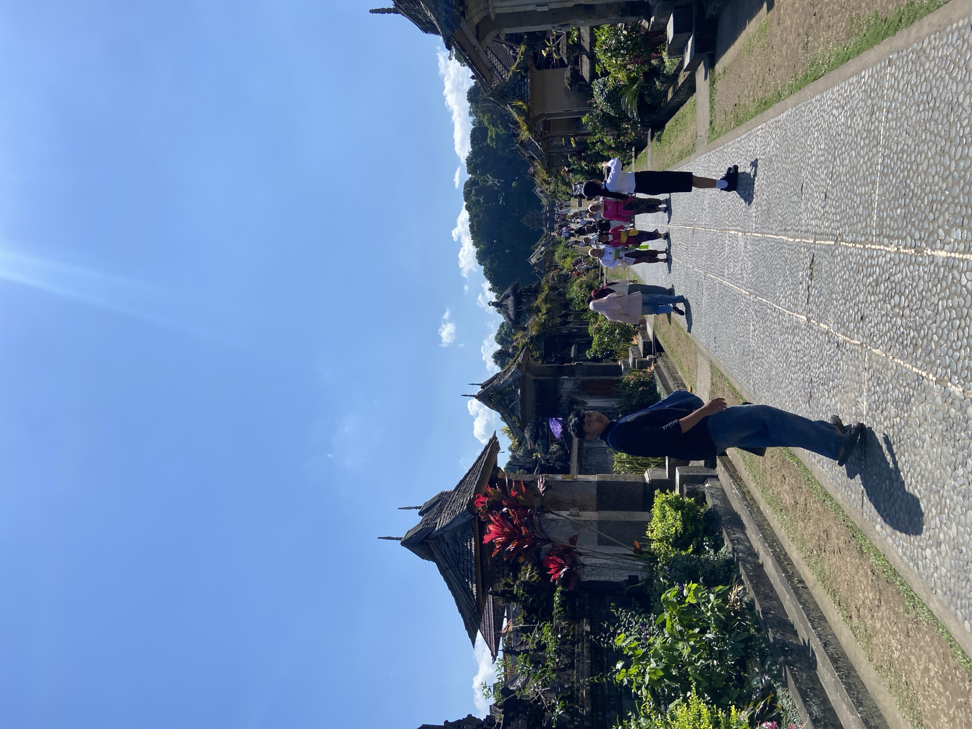 This is a full, eye-level shot of a pathway in an outdoor historical village, likely in Bangli, Indonesia, under a bright, sunny sky with scattered clouds. The pathway is paved with grey cobblestones and lined with traditional Balinese architecture featuring thatched roofs and intricate stone carvings. Lush green foliage and vibrant red plants grow along the sides of the path, adding a touch of color to the scene.
A diverse group of people, dressed in a mix of traditional and modern attire, are walking along the pathway, many appearing to be tourists. In the foreground, a young man in a dark shirt and blue jeans stands facing left, with a blue bag slung over his shoulder. He is the closest person to the camera and appears to be looking towards the viewer. Further down the path, several women in colorful outfits, including headscarves, are walking together. A young woman in a white shirt and black skirt is also visible, holding up a phone, possibly taking a photo or video. The pathway leads towards a densely wooded hill in the distance, suggesting a natural, picturesque setting for the village. The overall atmosphere is one of cultural exploration and tourism.