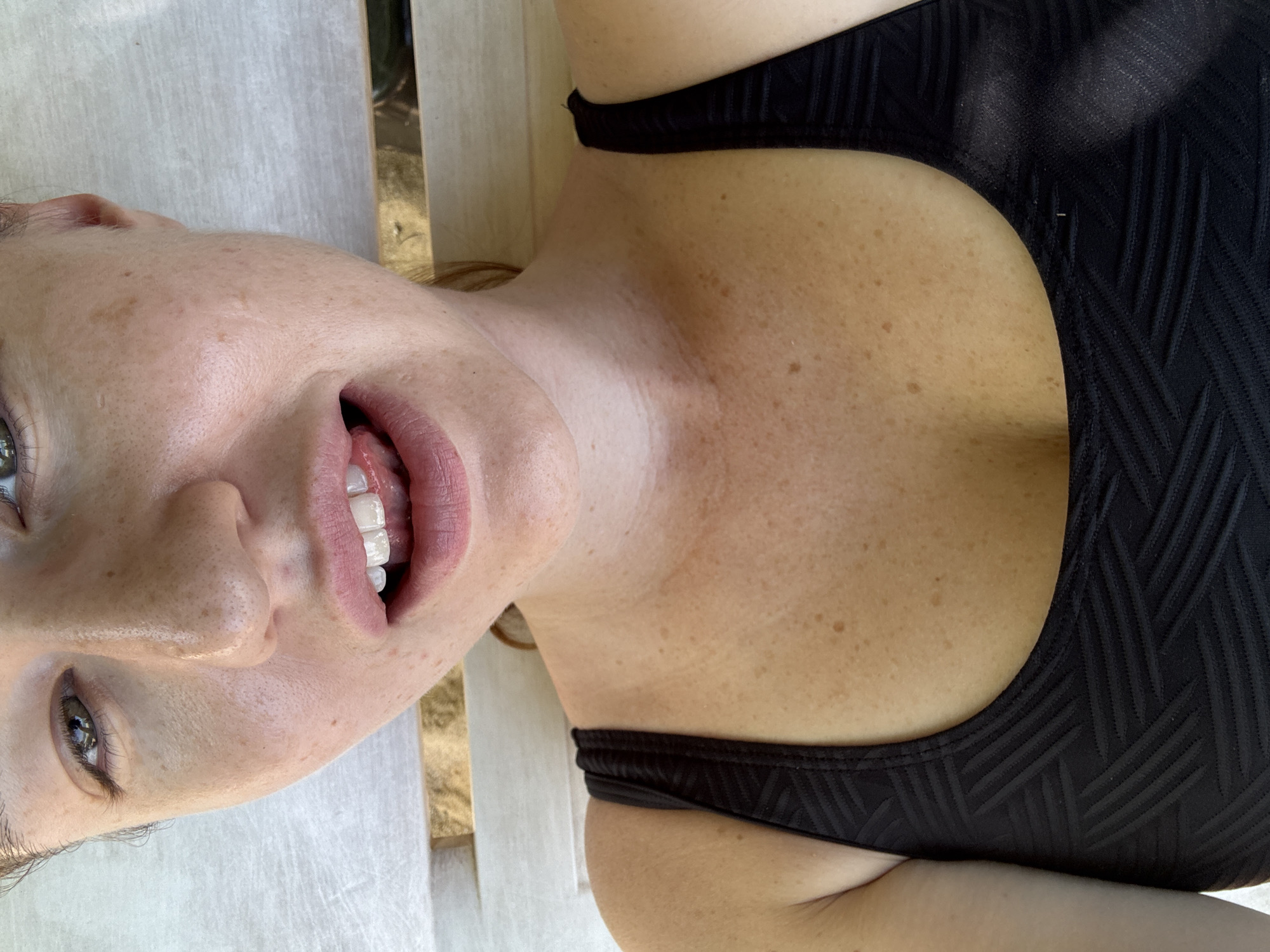 A close-up, overhead shot shows the upper torso and face of a person wearing a black patterned swimsuit. The person is lying down on a light-colored surface, possibly a lounger or deck chair, with wooden slats visible in the background, suggesting an outdoor setting in Rawai, Thailand. The person's face is partially visible, with their mouth open and tongue touching their upper teeth. Freckles are visible on their nose, cheeks, and chest. The lighting suggests it is daytime.