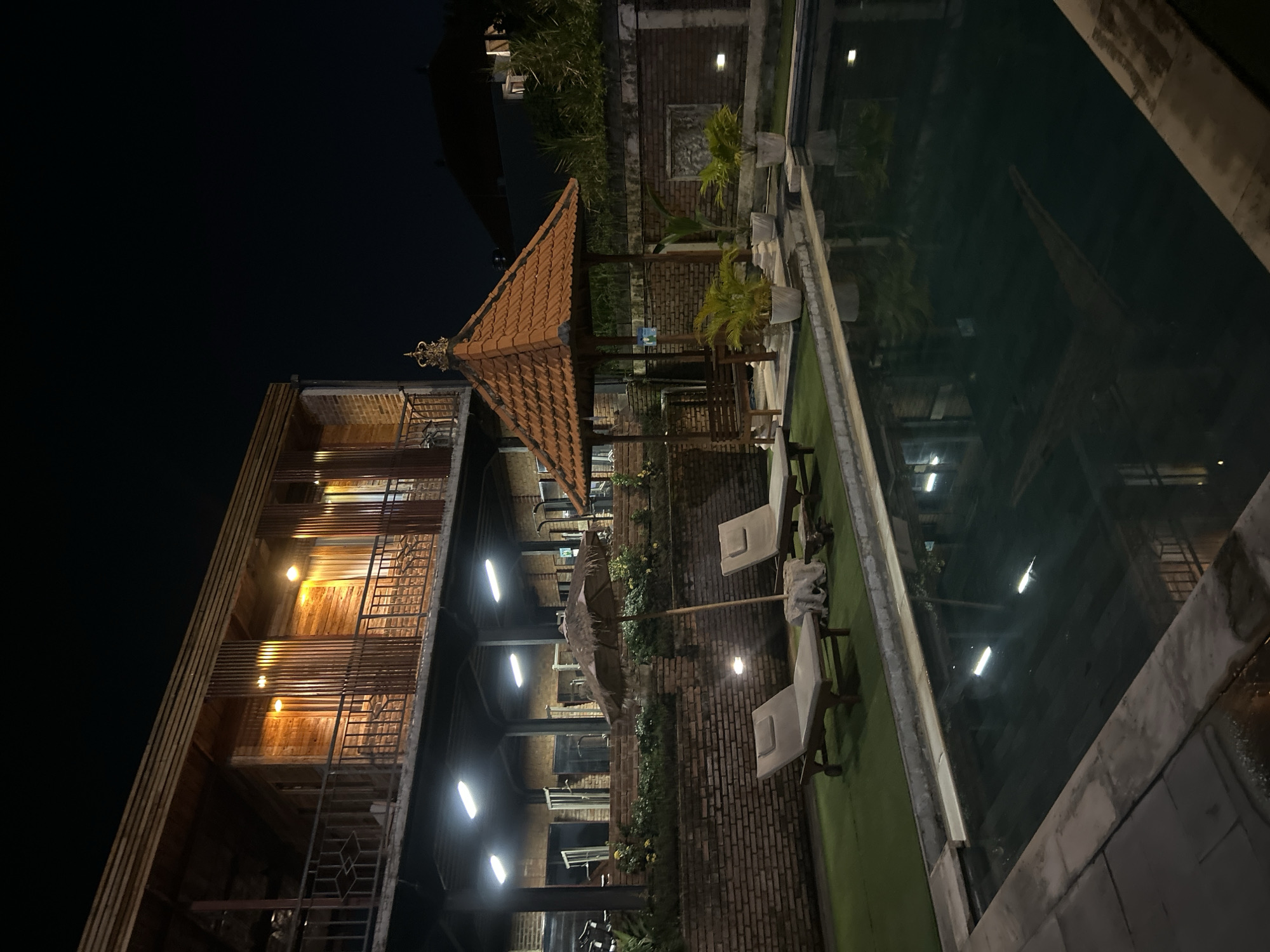 The image depicts a resort swimming pool area at night in North Kuta, Indonesia. The scene is illuminated by artificial lighting, casting a warm glow on the wooden architecture of a two-story building in the background. The building features balconies with railings and wooden slatted elements. In the foreground, the dark, reflective surface of a swimming pool occupies a significant portion of the frame.
Adjacent to the pool is a green, lawn-like area where two lounge chairs are placed under a thatched umbrella. A small wooden table sits between the chairs. Further to the right, a traditional gazebo with an orange tiled roof and decorative finial is visible. Several potted plants, including palm trees, are arranged around the gazebo and along the brick wall that encloses the area. The brick wall is a prominent feature, extending along the perimeter of the pool area. The overall atmosphere suggests a tranquil, tropical nighttime setting. There are no people visible in the image.