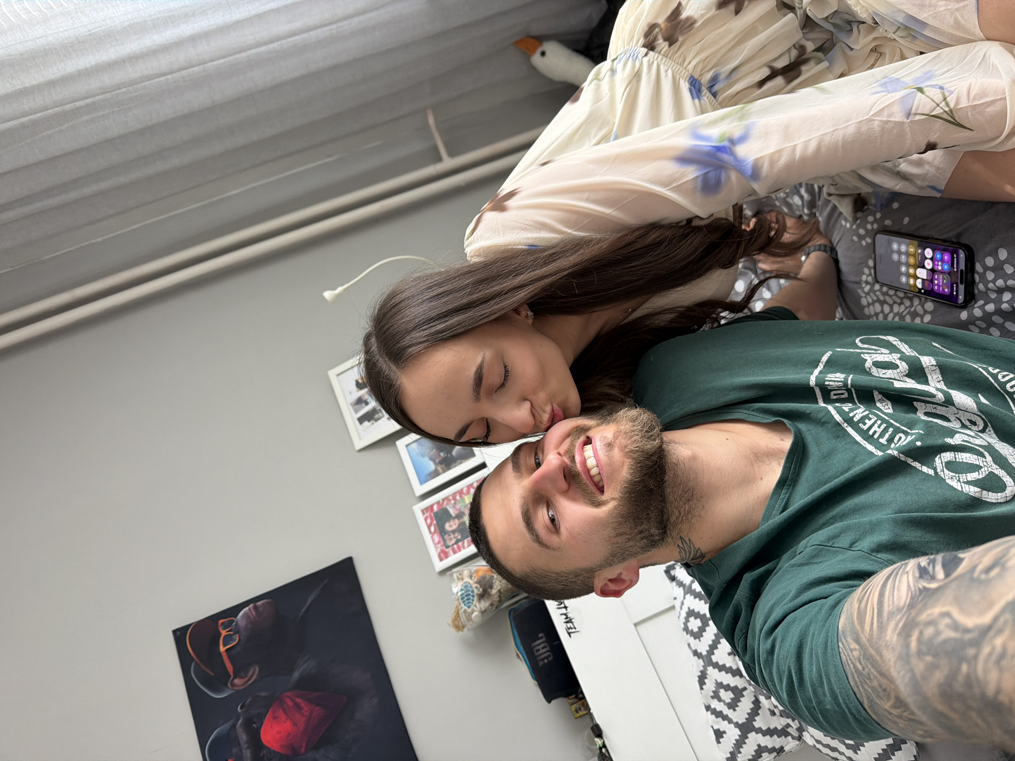 A couple is photographed indoors. The woman, with long dark hair, is leaning down, as if kissing the man on the cheek. The man has a beard and is wearing a green t-shirt. He is smiling and has tattoos on his arm. The background includes a wall with framed pictures, a JBL speaker, and an image of a figure in a black suit with red accents. A white and beige floral dress is also visible. A mobile phone is lying on the bed. The location is in Sofia, Bulgaria.
