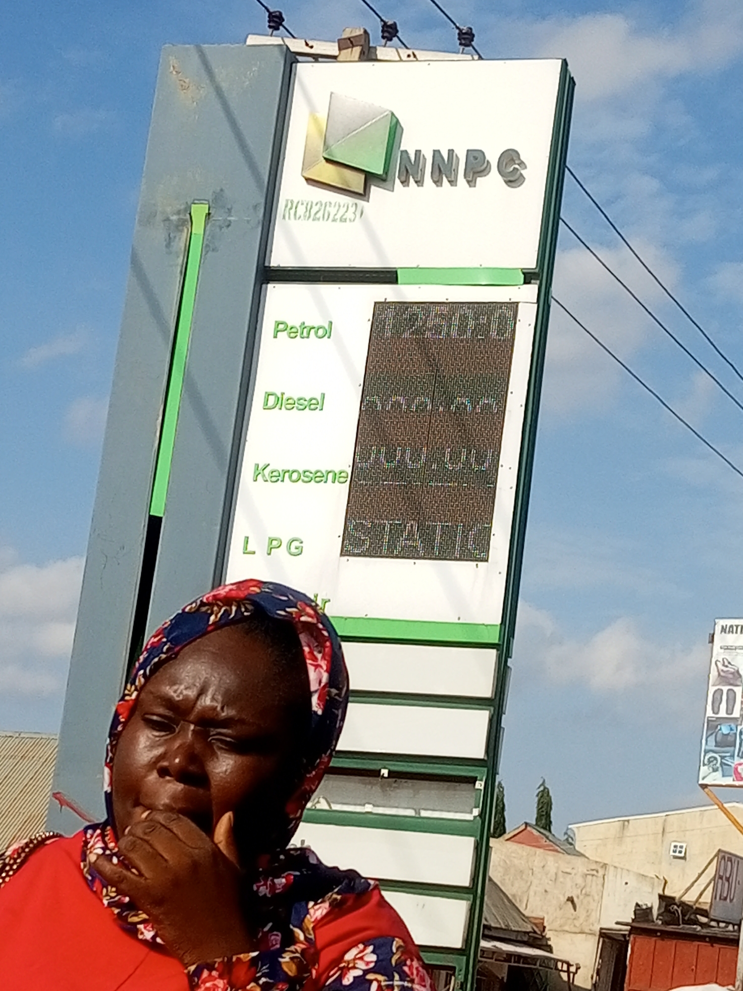 The image shows a woman in the foreground, wearing a red top and a headscarf with a floral pattern. She appears to be looking downwards with a pensive expression, her hand near her mouth. Behind her, a tall petrol station price board stands prominently. The board features the "NNPC" logo and lists fuel prices for "Petrol," "Diesel," "Kerosene," and "LPG" on digital displays. The prices for Petrol and Diesel are partially visible, showing "1250.00" and "300.00" respectively. The background reveals a blue sky with some clouds, suggesting it is daytime. The setting appears to be an outdoor location in Gombe, Nigeria, possibly a street or a commercial area, with hints of buildings and structures in the background. The overall mood is somewhat somber, conveyed by the woman's expression and the stark display of fuel prices.