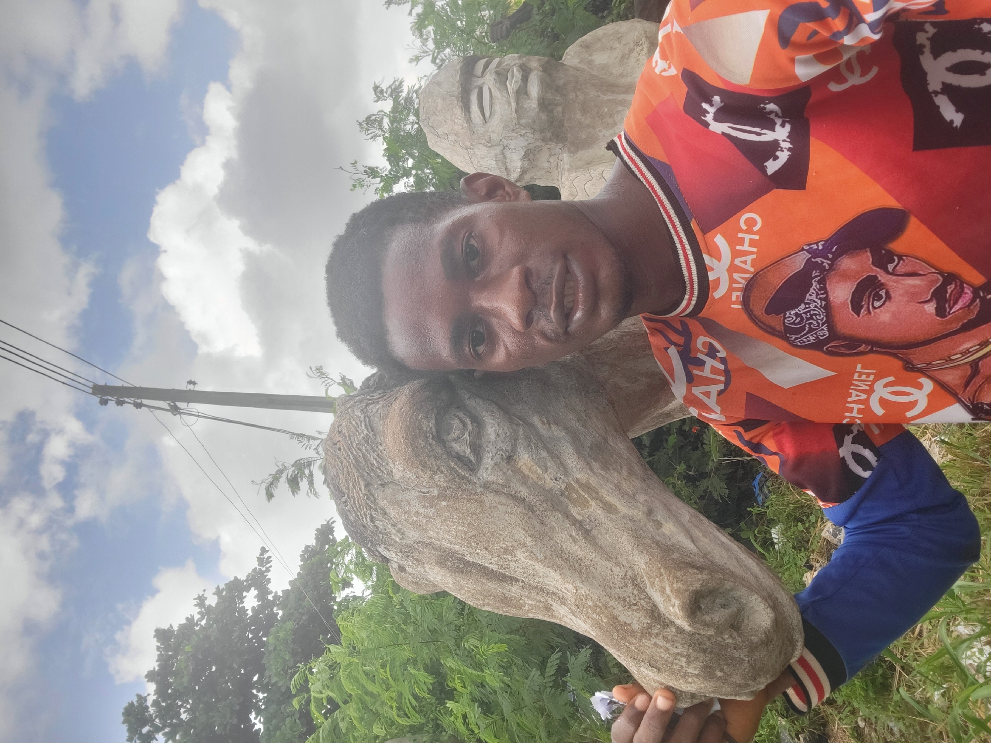 A young Black man is taking a selfie with a concrete sculpture of a horse's head in the foreground. He is wearing an orange and blue long-sleeved shirt with a repeating pattern of the Chanel logo and a portrait of Tupac Shakur. The sculpture is textured and weathered, with visible cracks. In the background, out of focus, is another concrete sculpture of a human head with closed eyes. The scene appears to be outdoors, with lush green foliage and a utility pole with wires visible in the upper right corner. The sky is partly cloudy with patches of blue. The overall setting is consistent with an outdoor art installation or public space in Egbagu, Nigeria.