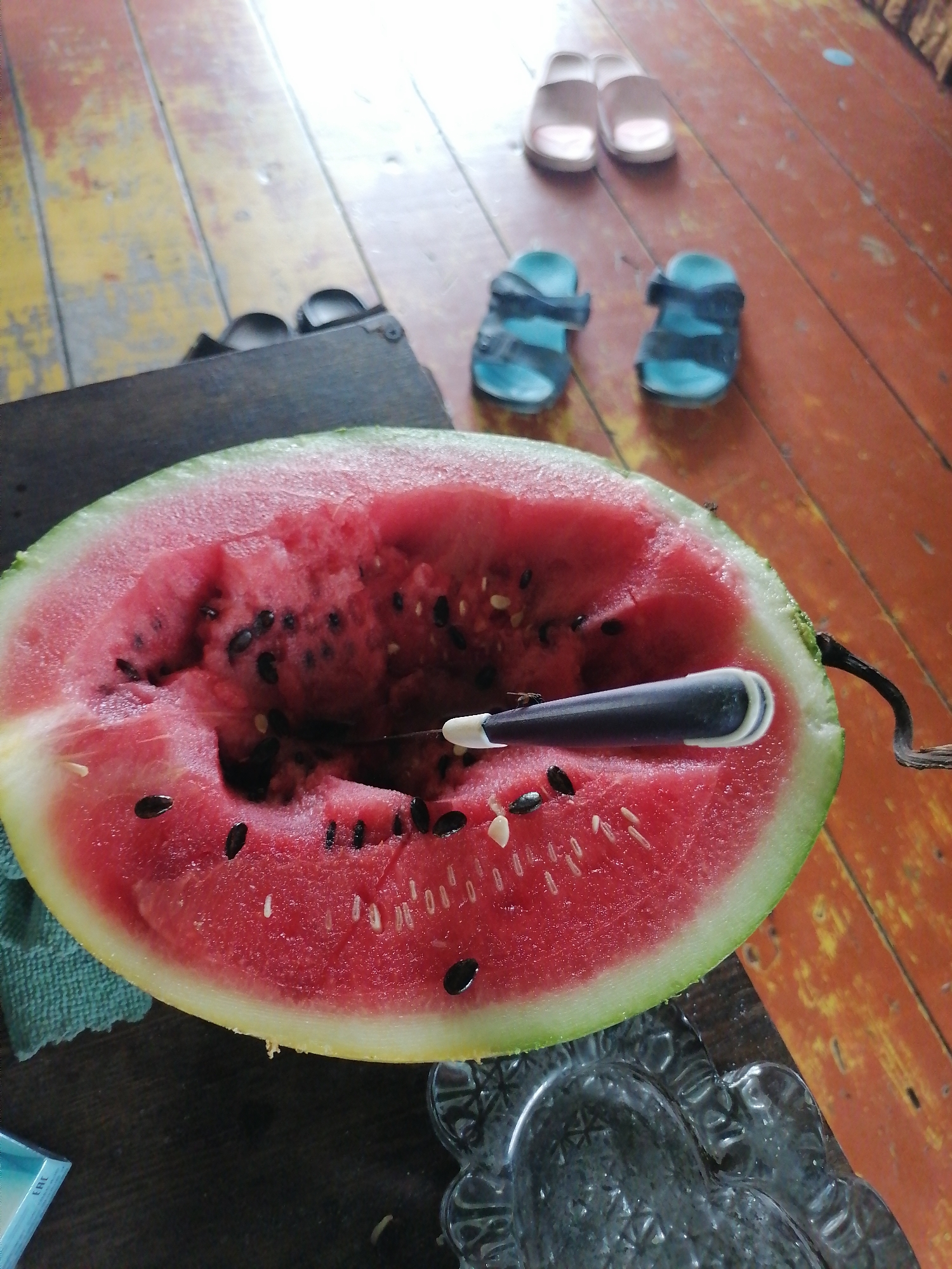 A half-eaten watermelon with black seeds and some white seeds is on a wooden surface. A dark-handled utensil with a white tip is inserted into the watermelon. The watermelon is on a teal cloth. In the background, two pairs of sandals are on the wooden floor, and a dark rectangular object is to the left of the watermelon. The floor is made of weathered orange and yellow planks. The scene is indoors.
