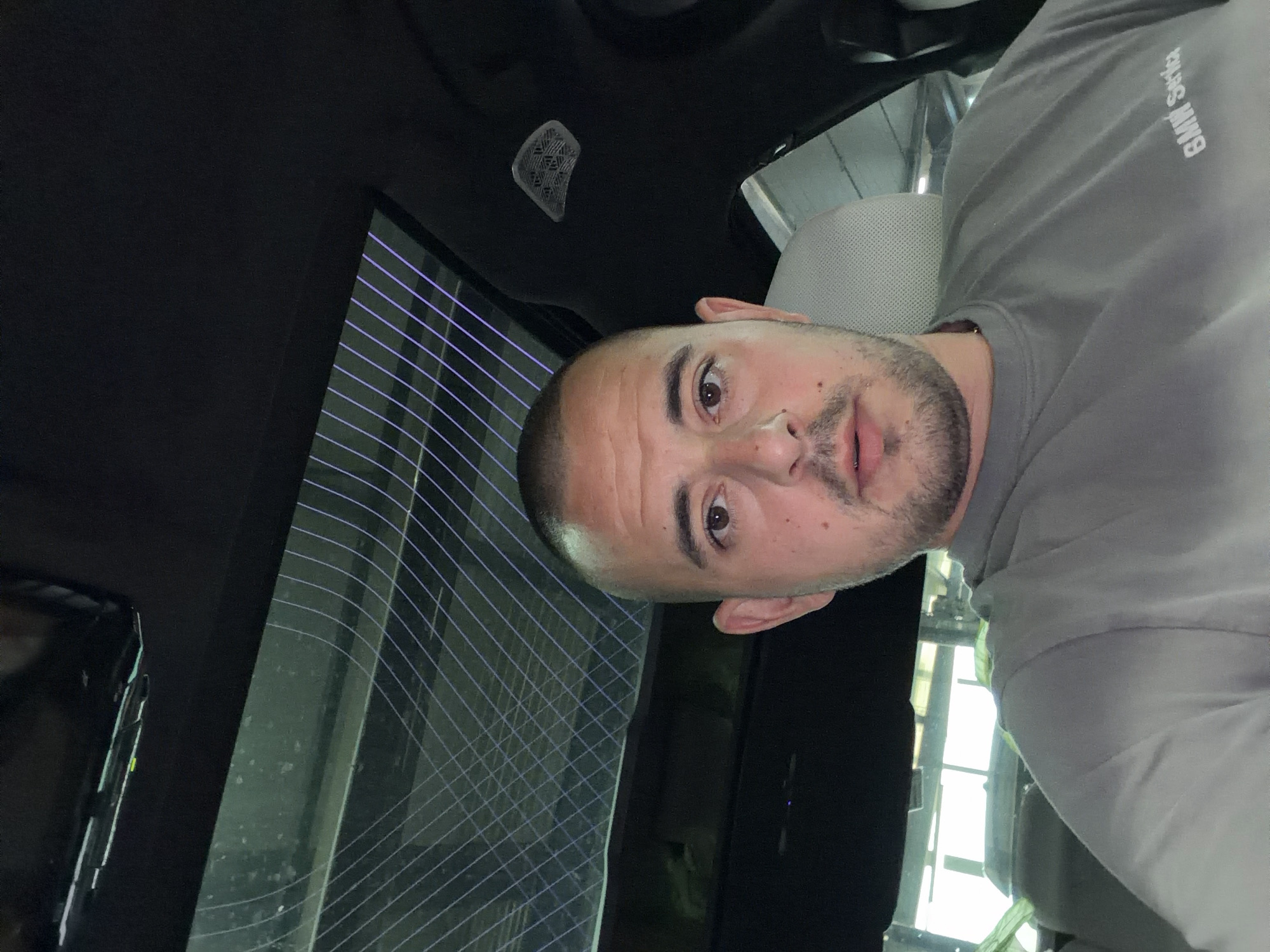 A man with a shaved head and a short beard is taking a selfie inside a car. He is wearing a grey t-shirt with "BMW Service" printed on the left chest area. The car has a panoramic sunroof with a grid pattern, and a light fixture is visible on the roof liner. The man is looking directly at the camera with a neutral expression. The background appears to be an indoor space, possibly a garage or workshop, with bright lights and some indistinct structures. The man's location is not specified in the provided image or crops.