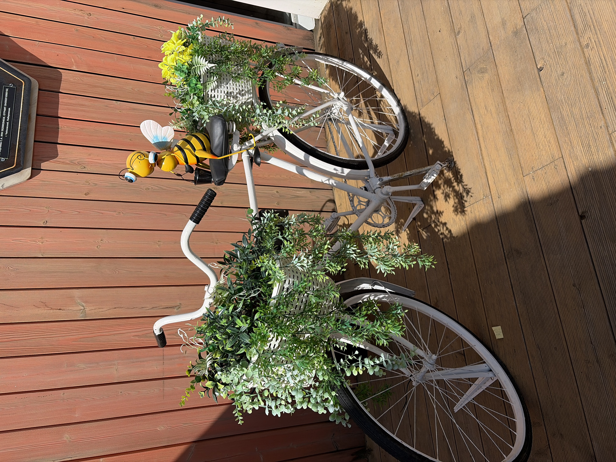 A tilted, medium shot presents a white bicycle decorated with plants and a bee ornament, situated on a wooden deck. The bicycle features a basket filled with greenery and yellow flowers on the front, and another basket overflowing with greenery on the rear. A yellow bee ornament is perched on the handlebars. The wooden deck consists of parallel planks, with varying shades suggesting different areas of sun exposure. The overall impression is of a decorative, repurposed bicycle adding a touch of whimsy to an outdoor space. No specific city or location is discernible.