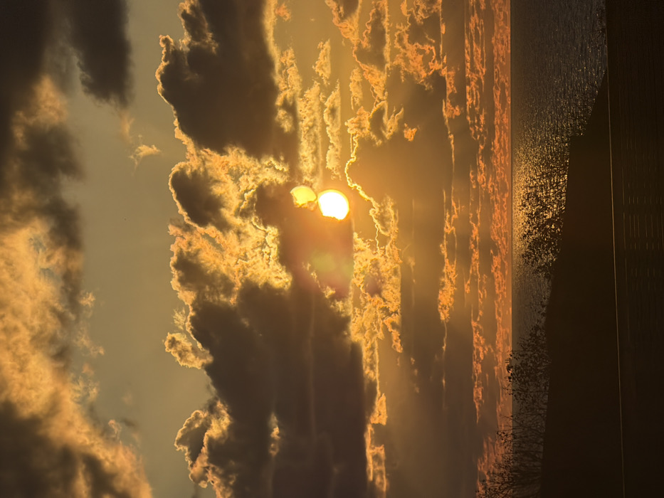 A mesmerizing sunset illuminates a sky filled with textured clouds. The sun, a radiant orb, shines through breaks in the cloud cover. Light beams and reflections dance on a body of water that stretches to the horizon. A dark, possibly silhouetted shoreline is visible on the right, marked by vegetation and what might be a building or structure. The overall palette is dominated by warm hues of orange and gold, contrasted by darker cloud formations.
