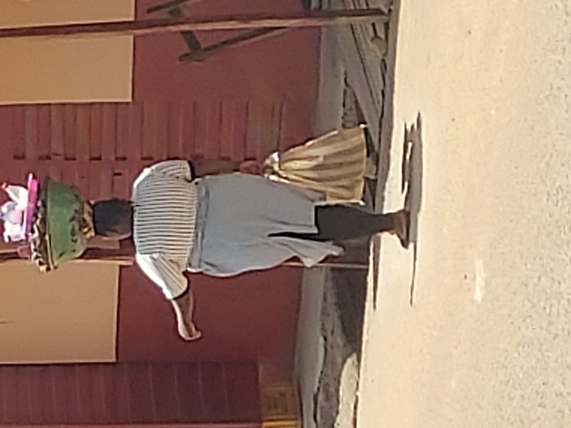 An elderly woman walks on a city street in Jos, Nigeria. She carries a basket with goods on her head and another object in her right hand. The area is urban, with a building visible in the background.
