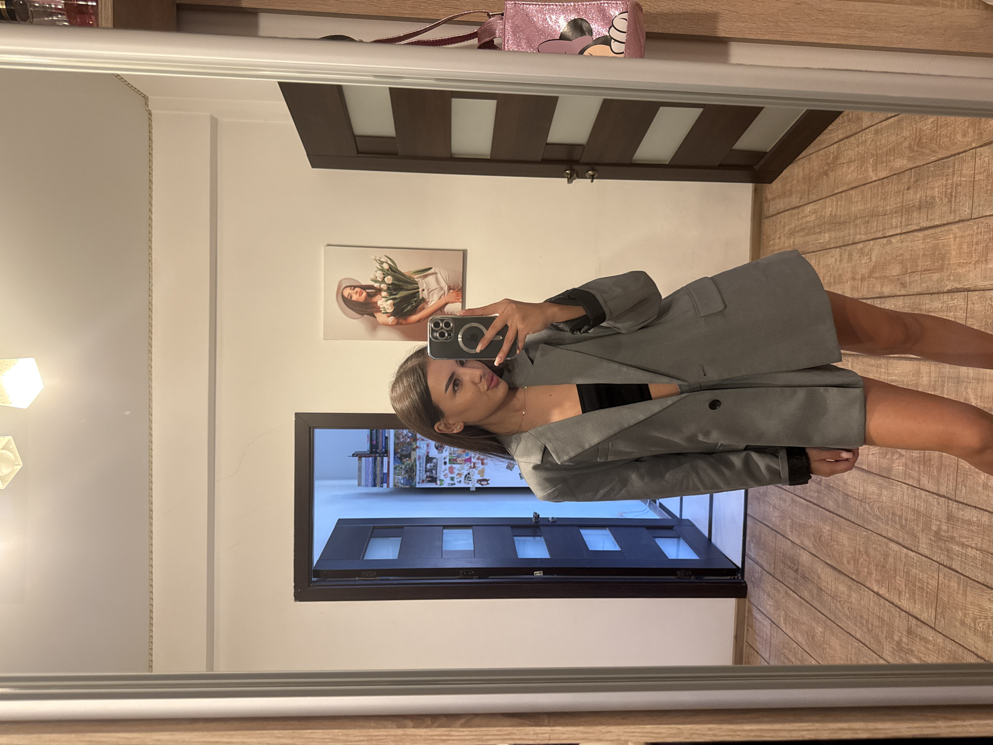 The image is a mirror selfie taken by a young woman indoors, possibly in an apartment in Chișinău, Moldova. She is wearing a gray oversized blazer over a black top and shorts. Her hair is dark brown and she is wearing subtle makeup. She is holding a smartphone, taking the picture. In the background, there is a door with a window, a picture on the wall of a woman holding tulips, and a pink sparkly bag with a cartoon character on it hanging on a shelf. The floor is made of wooden planks. The room appears to be a bedroom or dressing area.