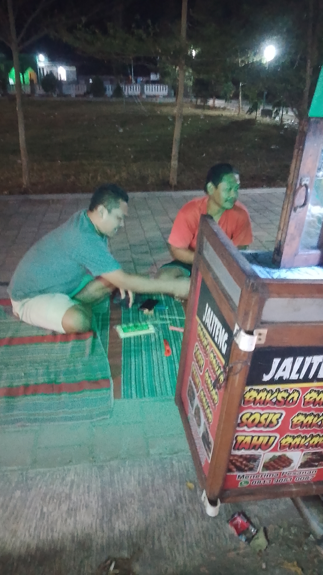 In Karanglangu, Indonesia, two men are seated on mats on a paved area at night. The man on the left, wearing a teal t-shirt and light-colored shorts, is crouching and appears to be interacting with items on the mat in front of him. To his right, a man in an orange t-shirt is sitting and looking towards his left. In the foreground, a food cart is positioned at an angle, displaying menus with Indonesian text and images of food items. The cart has a wooden frame and features signage that reads "JALITENG" and lists items such as "BAKSO", "SOSIS", and "TAHU". Beneath these item names, a phone number "0813 9081 0087" is visible, along with the text "Menerima Pesanan" (Taking Orders). The background shows trees, a building with illuminated windows, and what appears to be a grassy area, suggesting an outdoor setting, possibly a park or public space at night.