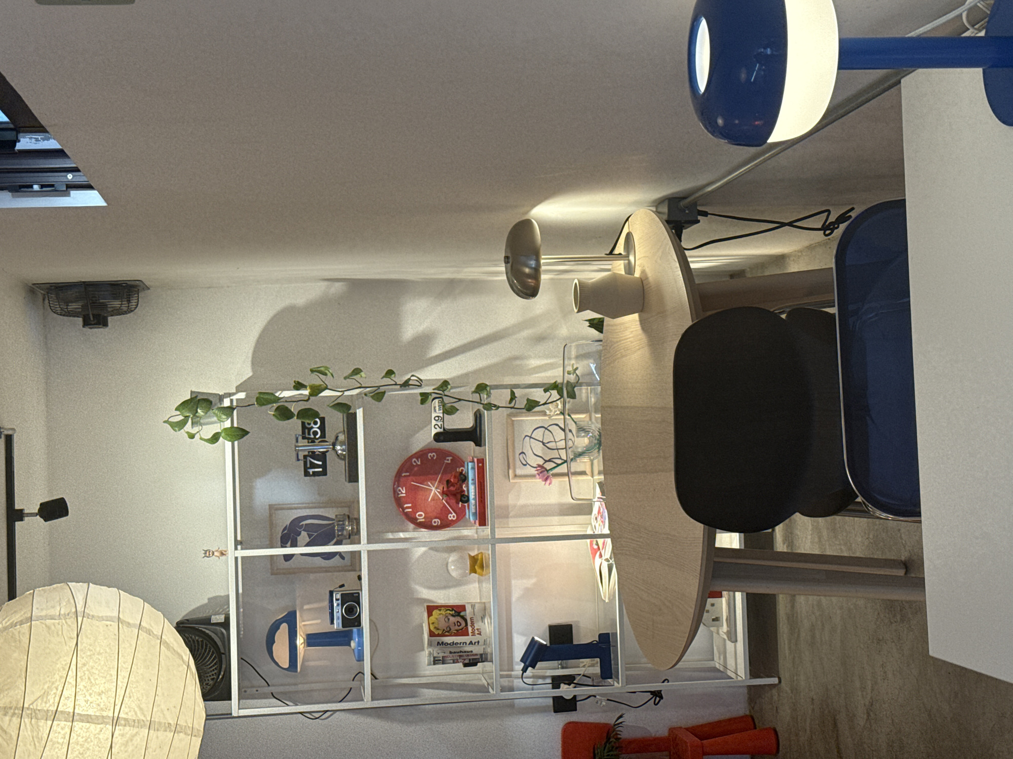 This image presents a modern and eclectic indoor space, possibly a living area or studio, with a focus on organized shelves and unique lighting. The room is brightly lit, primarily by artificial light sources.
On the left, a large, spherical white paper lantern rests on the floor next to a black box fan. Above it, a black wall-mounted lamp extends into the space. A metal exhaust fan is visible on the ceiling in the upper left corner.
The central feature is a white, open modular shelving unit. The top shelf displays a green plant vine, a digital flip clock showing "17 58" (5:58 PM), a small date indicator reading "29 WED", a red circular wall clock (indicating approximately 10:07), and a small framed drawing. The middle shelf contains a framed blue abstract artwork and a vintage-style camera. The bottom shelf holds several books, including spines with the text "Modern Art" and "Bauhaus," alongside a blue directional lamp.
To the right, a light-colored wooden table with an irregularly shaped top is flanked by two chairs featuring dark seats and striking blue backrests. Overhead, a distinctive blue and white spherical ceiling lamp is visible. Exposed metal conduits run along the ceiling. Red plastic objects are partially visible on the floor in the bottom right corner.
The time displayed on the flip clock suggests late afternoon or early evening. The space appears tidy and curated, combining functional elements with artistic decor. No people are present, and no specific activity is taking place. While the location context is San Andrés Cholula, Mexico, there are no direct visual cues within the image to confirm this.